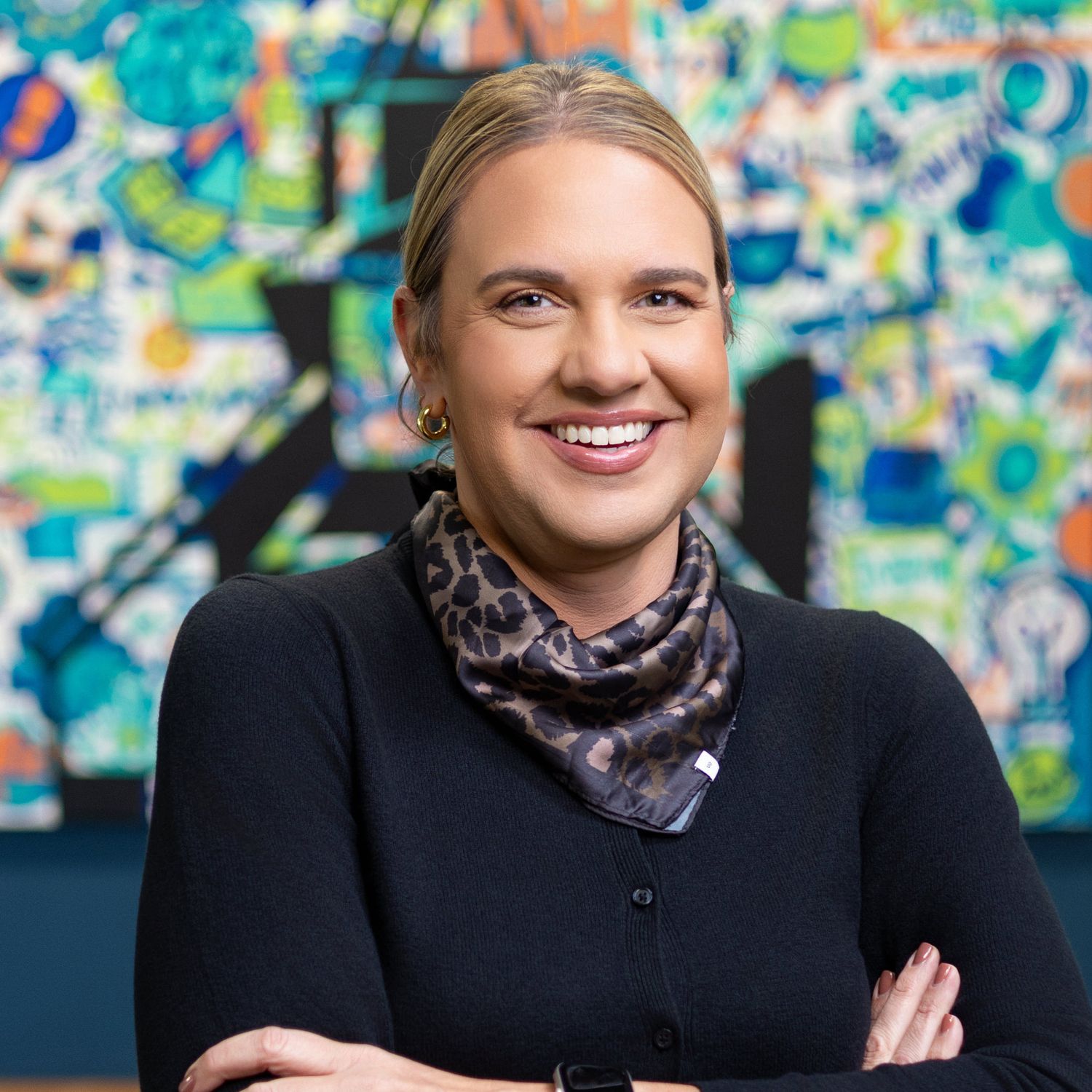 Professional portrait of someone in a black sweater and patterned scarf standing with crossed arms against colorful artwork.