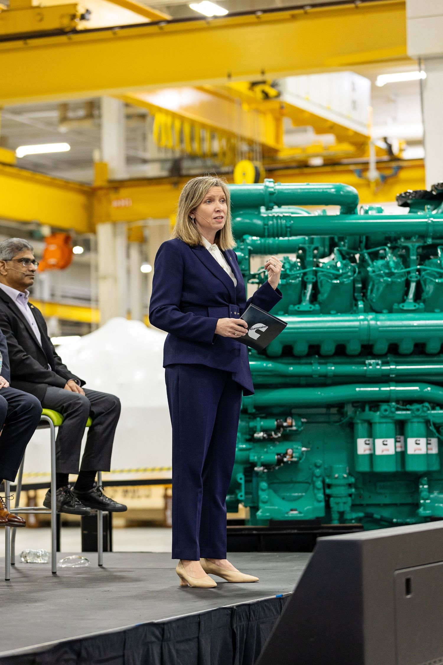 Professional business presentation with a speaker standing next to a large green industrial engine in a factory setting.
