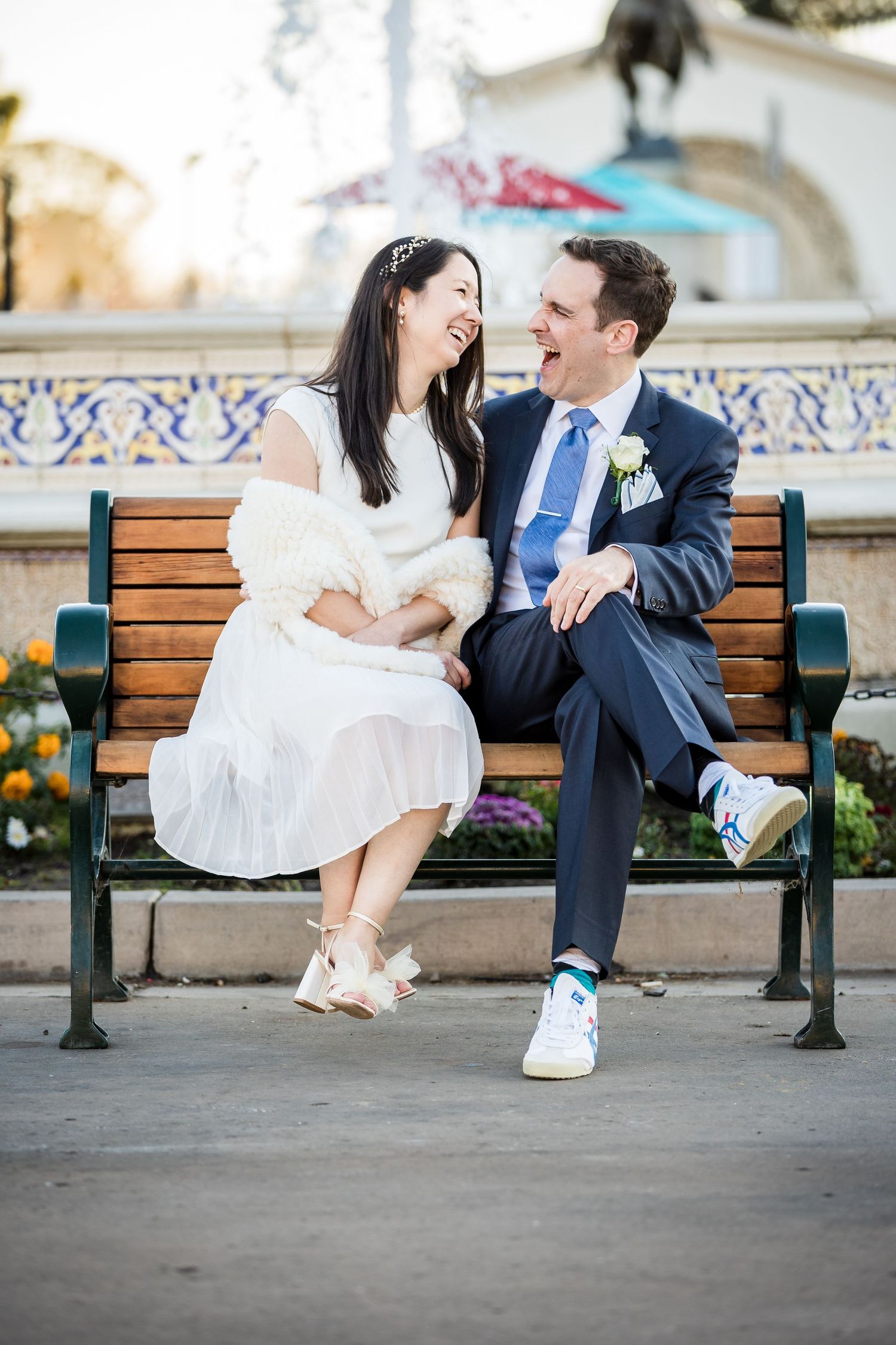 A couple shares a joyful moment sitting on a park bench wearing wedding attire with contrasting formal and casual shoes.