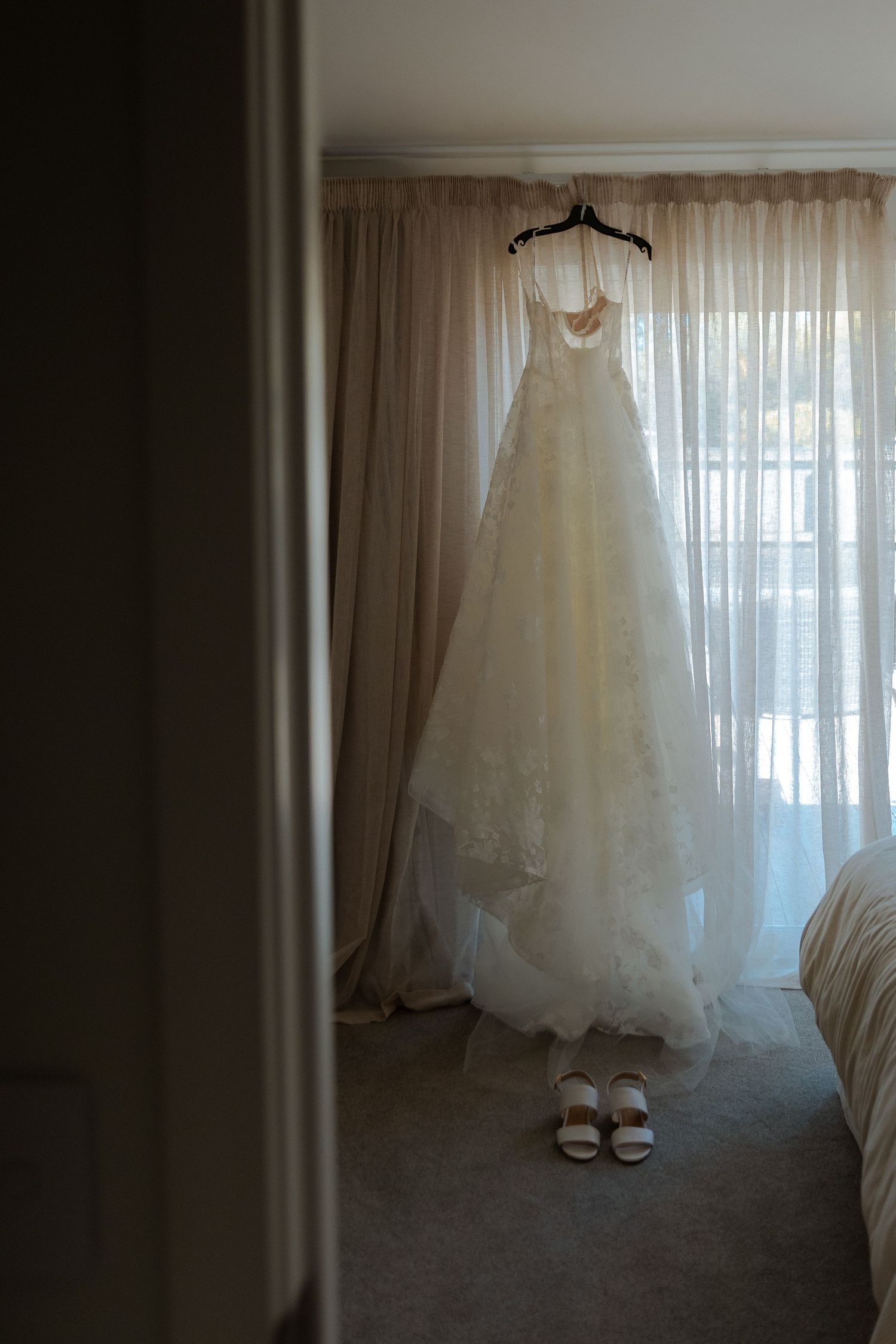Elegant white lace wedding dress hanging on a hanger in front of sheer curtains with bridal sandals below.