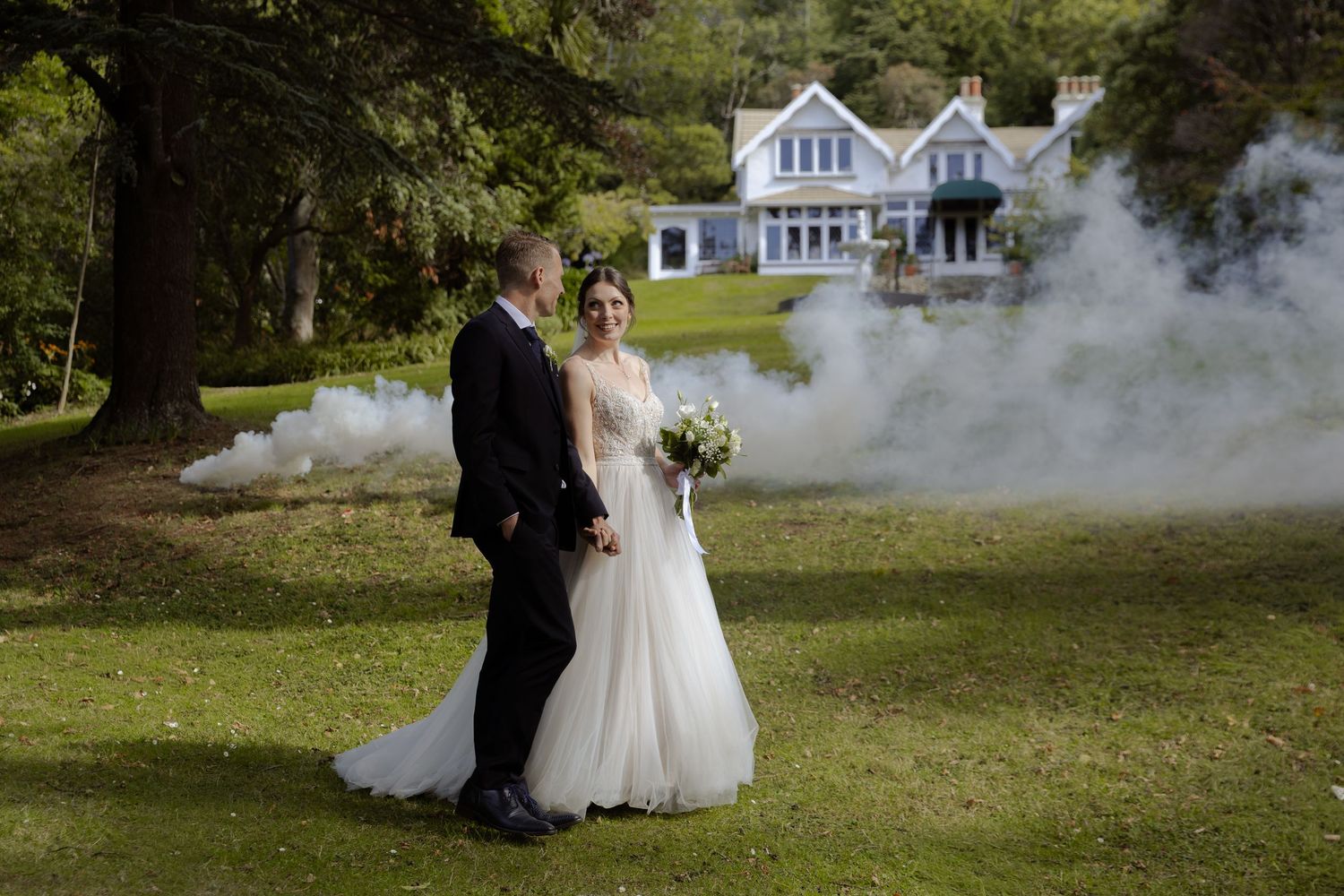 Bride and groom pose on lush green lawn with dramatic smoke effect and elegant manor house backdrop.