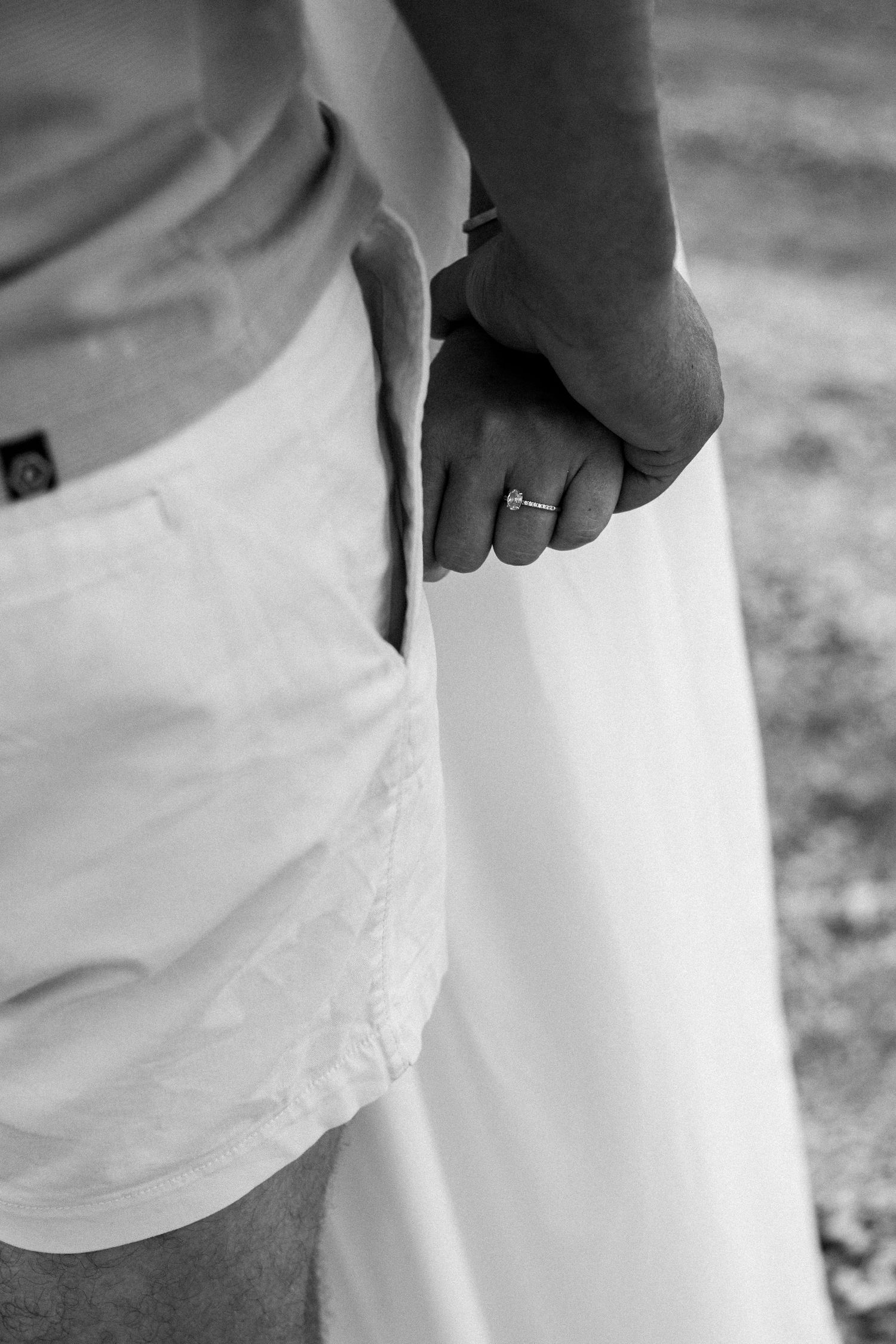 Black and white photo of a couple holding hands, bride wearing engagement ring, groom in white shorts.
