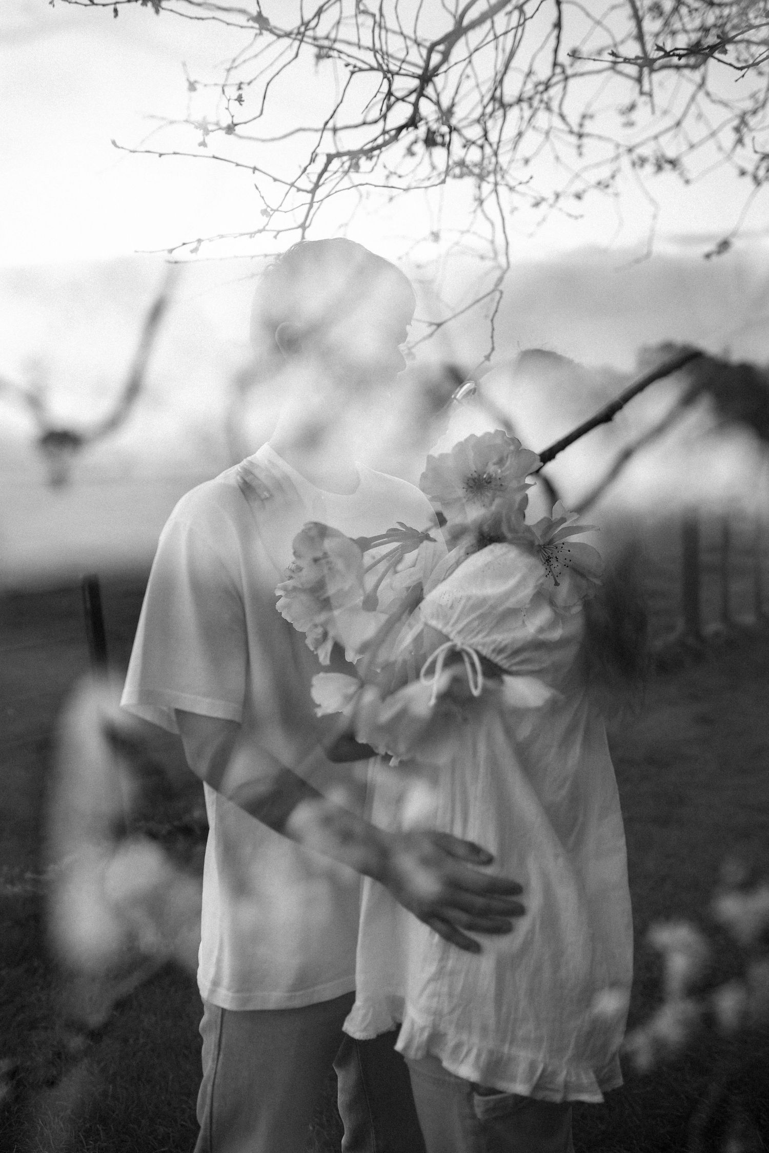 Black and white photo of a couple embracing outdoors, woman holding flowers, soft bokeh foreground, bare trees in background.