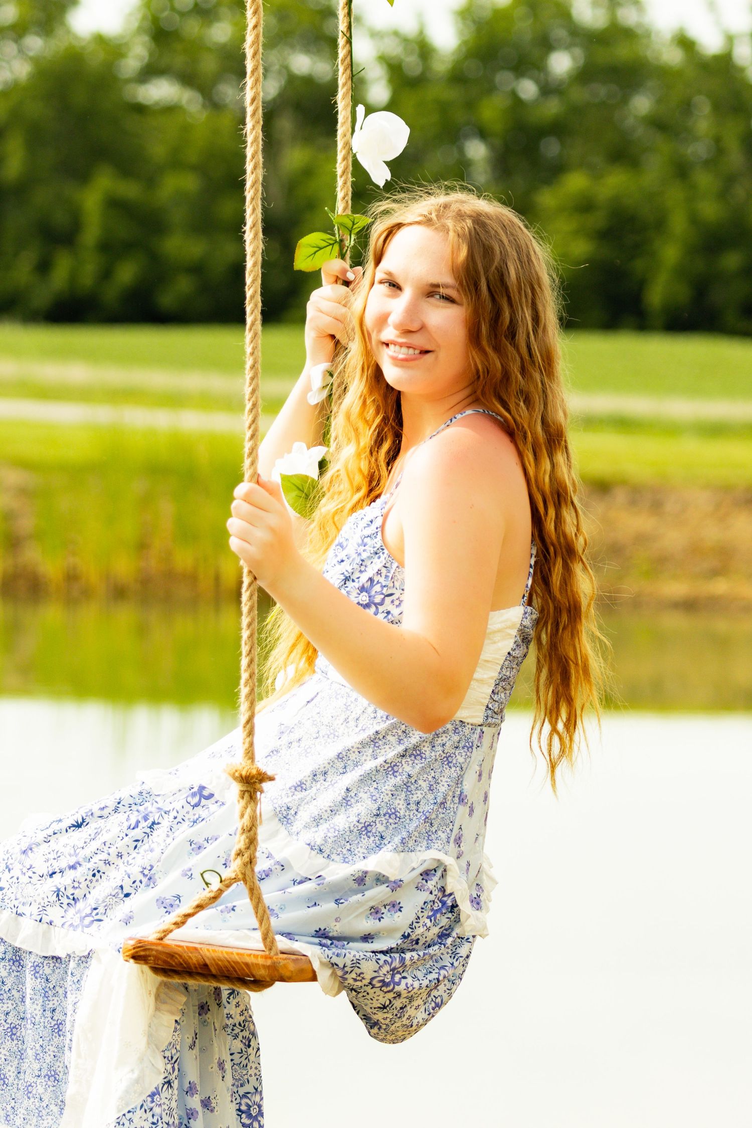 Young woman in floral dress sitting on rope swing holding white flower, smiling outdoors near pond.