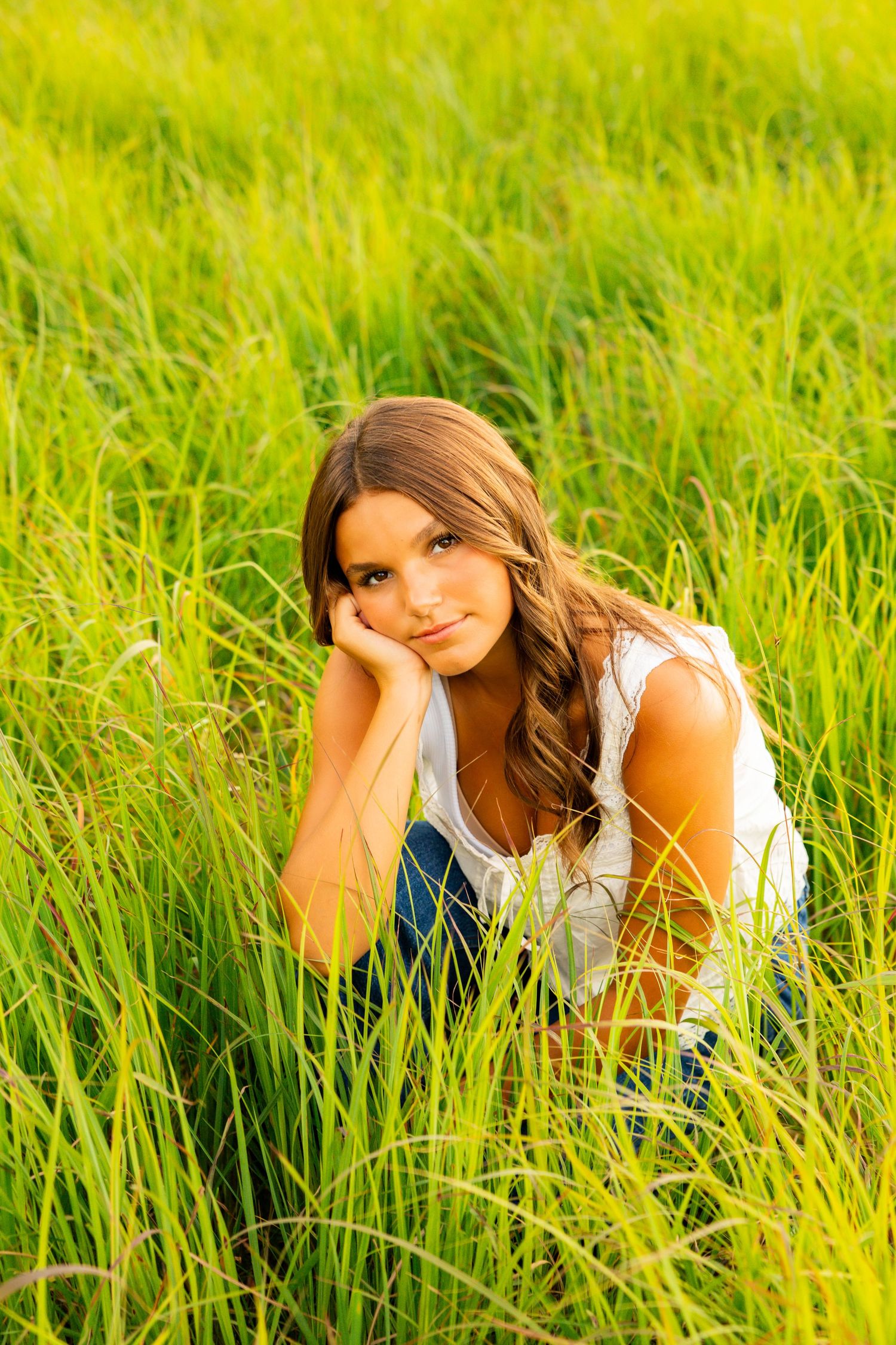 Young woman with wavy hair resting in tall green grass field, looking at camera in natural outdoor setting.