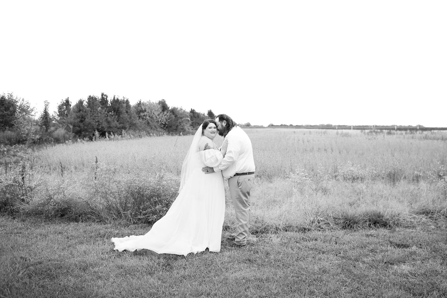A romantic black and white wedding photo of a couple embracing in a rustic field with trees in the background.