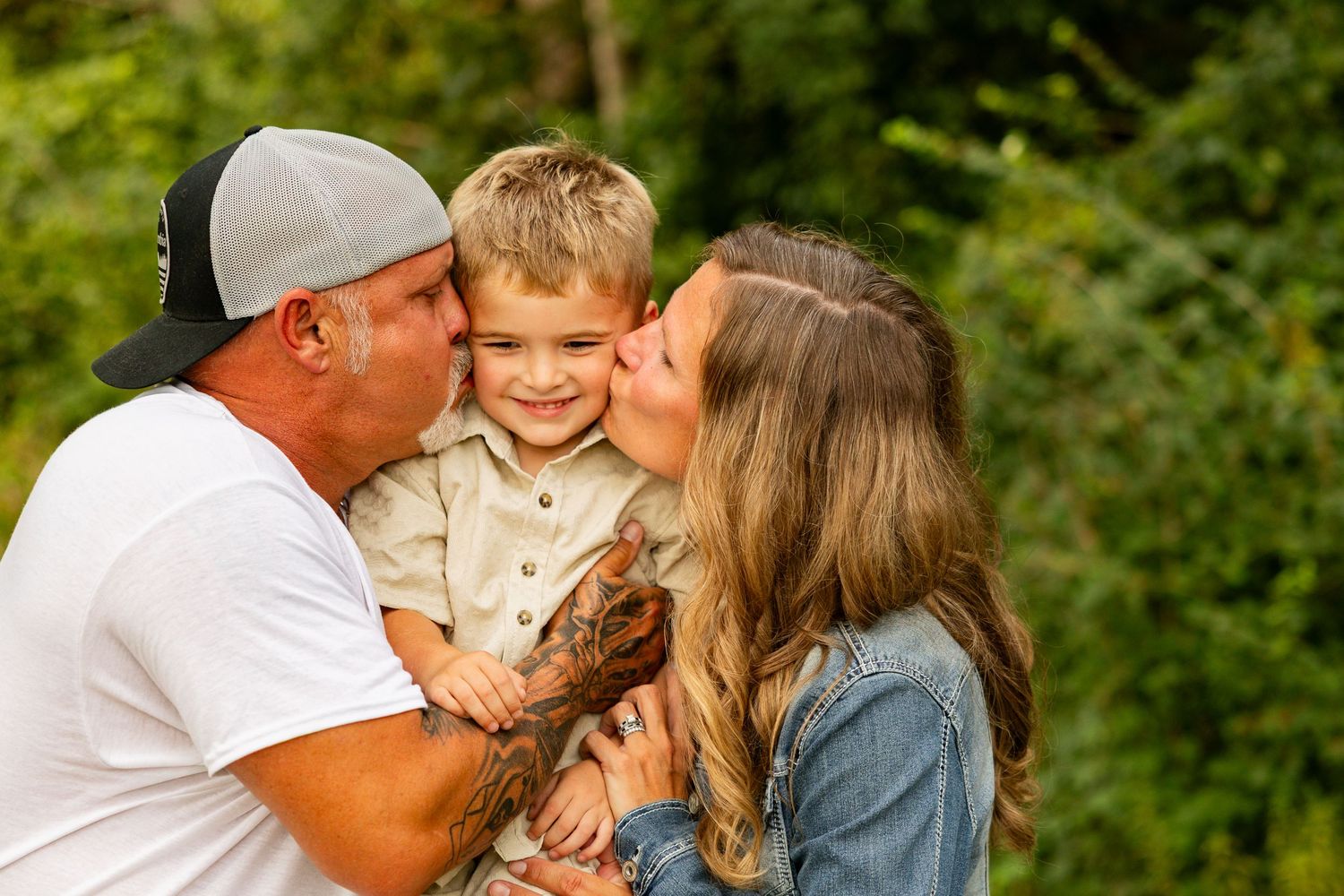 Parents kissing their smiling toddler boy on the cheeks outdoors with green trees in the background.