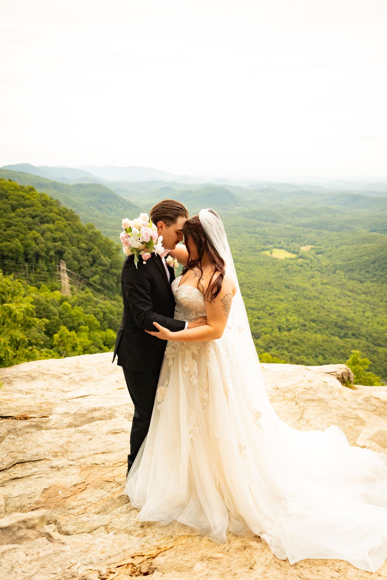 A romantic wedding couple embraces on a scenic mountain overlook with rolling green hills stretching into the distance.
