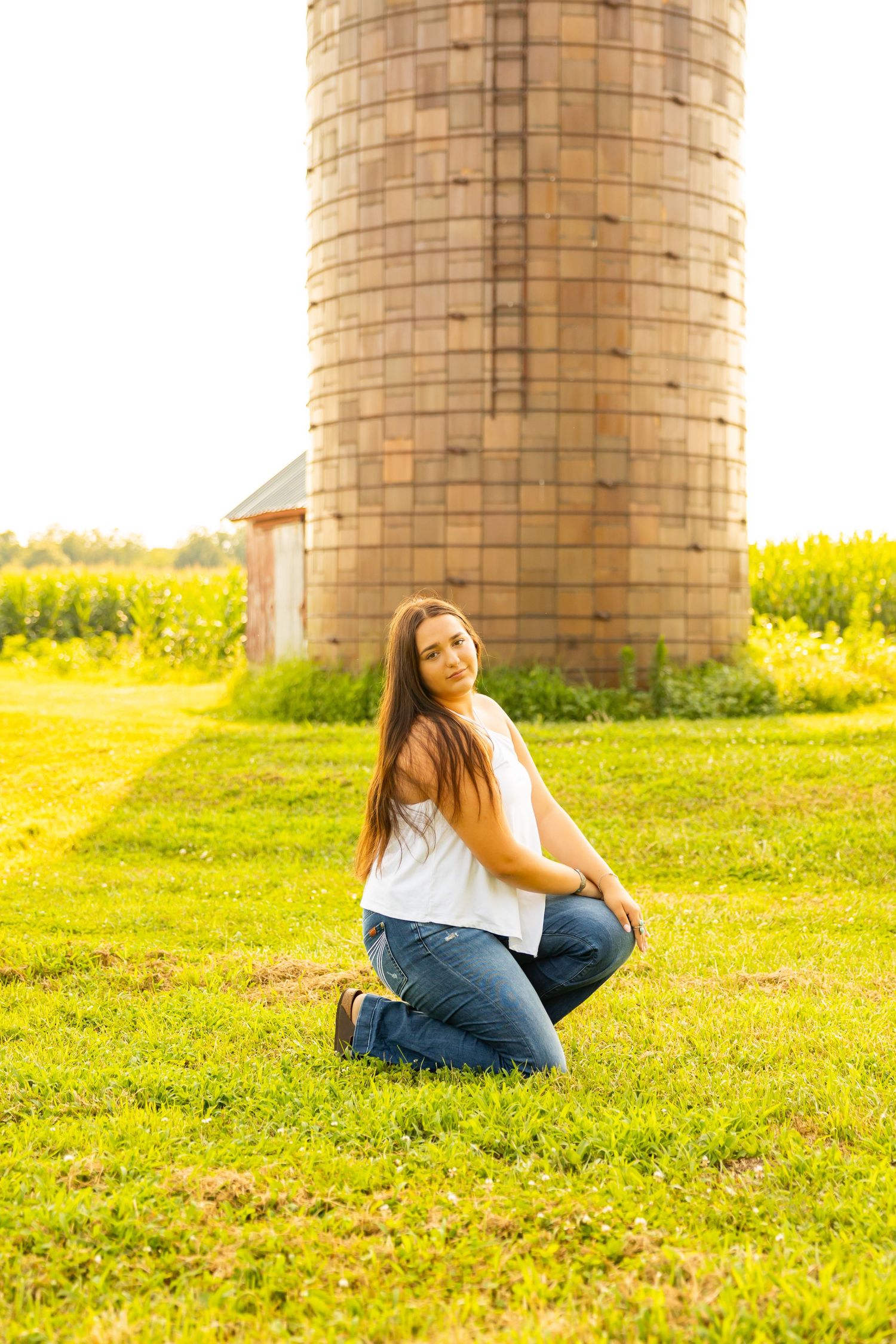 Young woman in white top and jeans kneeling on green grass in front of a rustic brick silo on a farm.