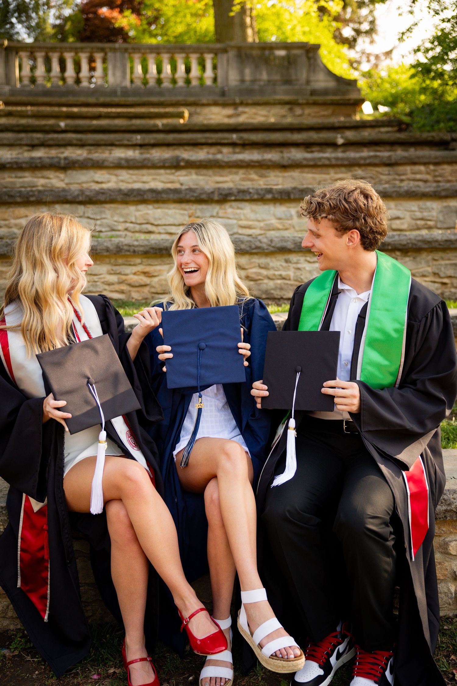 Graduates in black gowns and colorful stoles lean against stone wall in playful poses during graduation photo shoot.