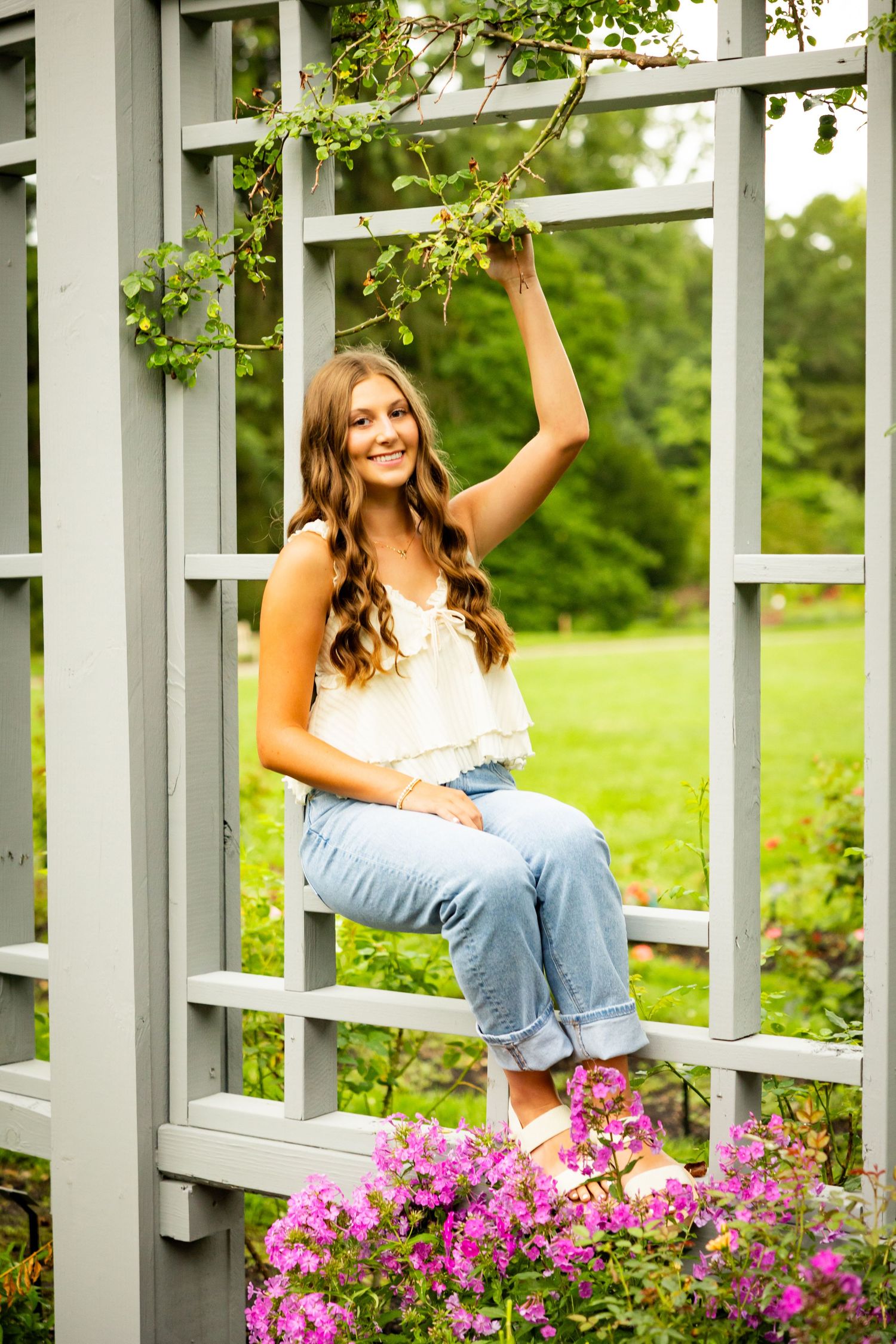 A person sits casually on a gray garden arbor with pink flowers blooming below on a sunny summer day.