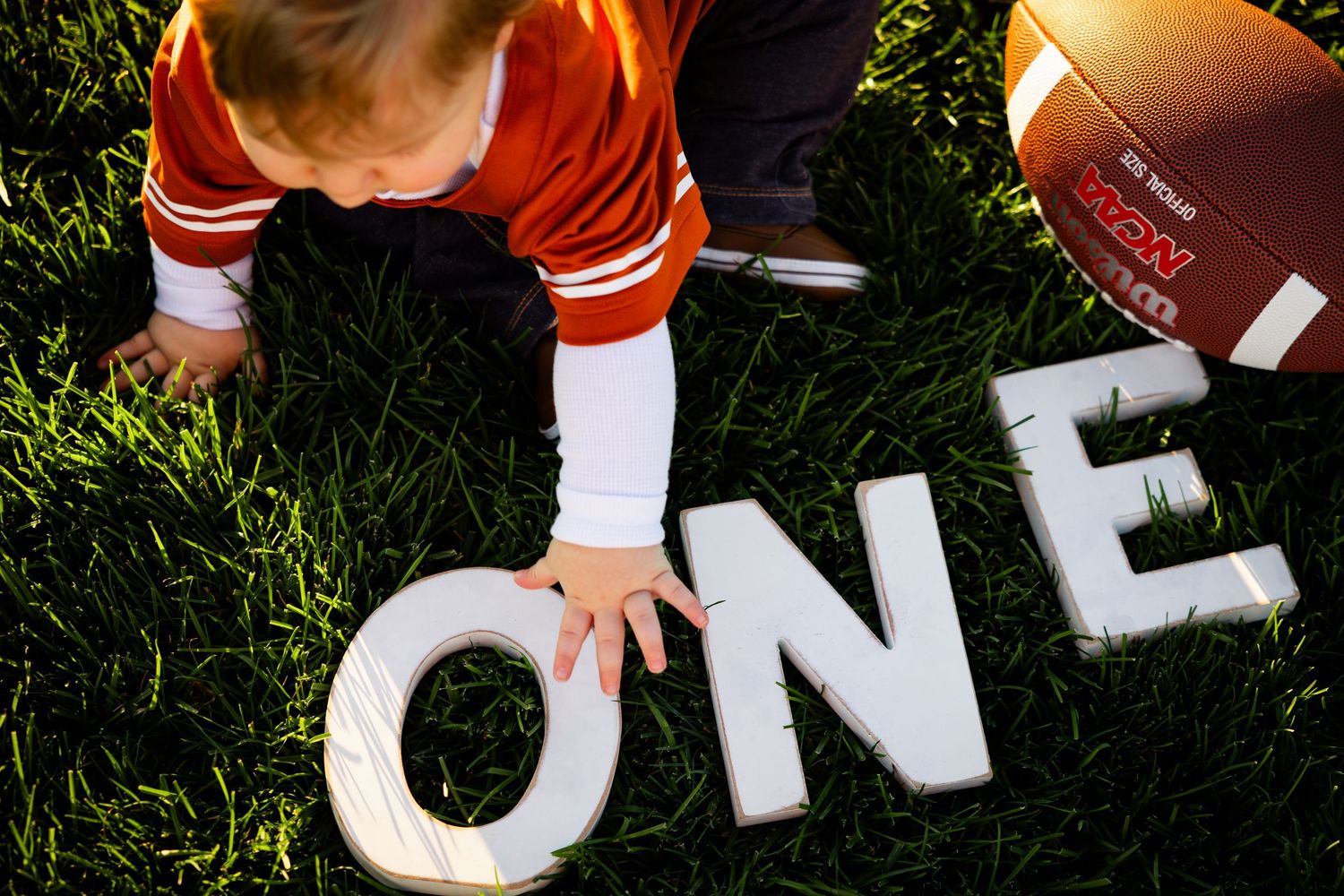 A small child touches white block letters spelling 'ONE' on grass next to a football.