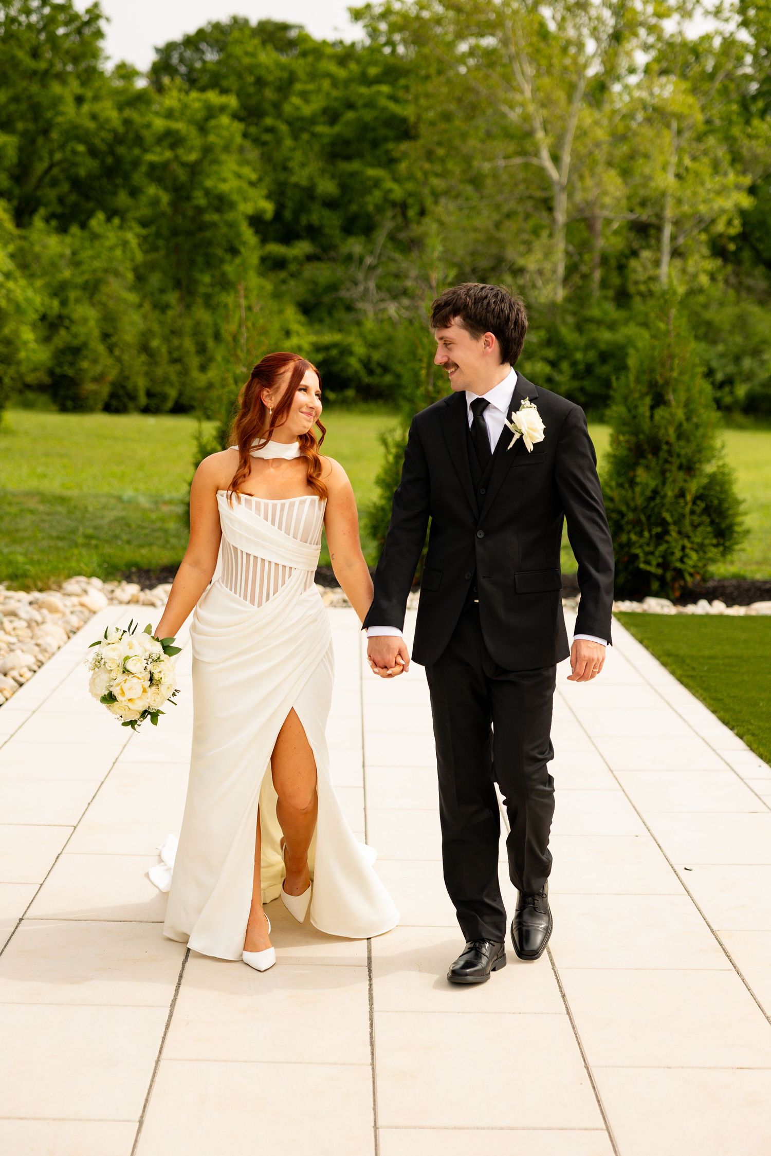 A bride in a white gown and groom in a black suit walk hand in hand on a stone path surrounded by lush greenery.