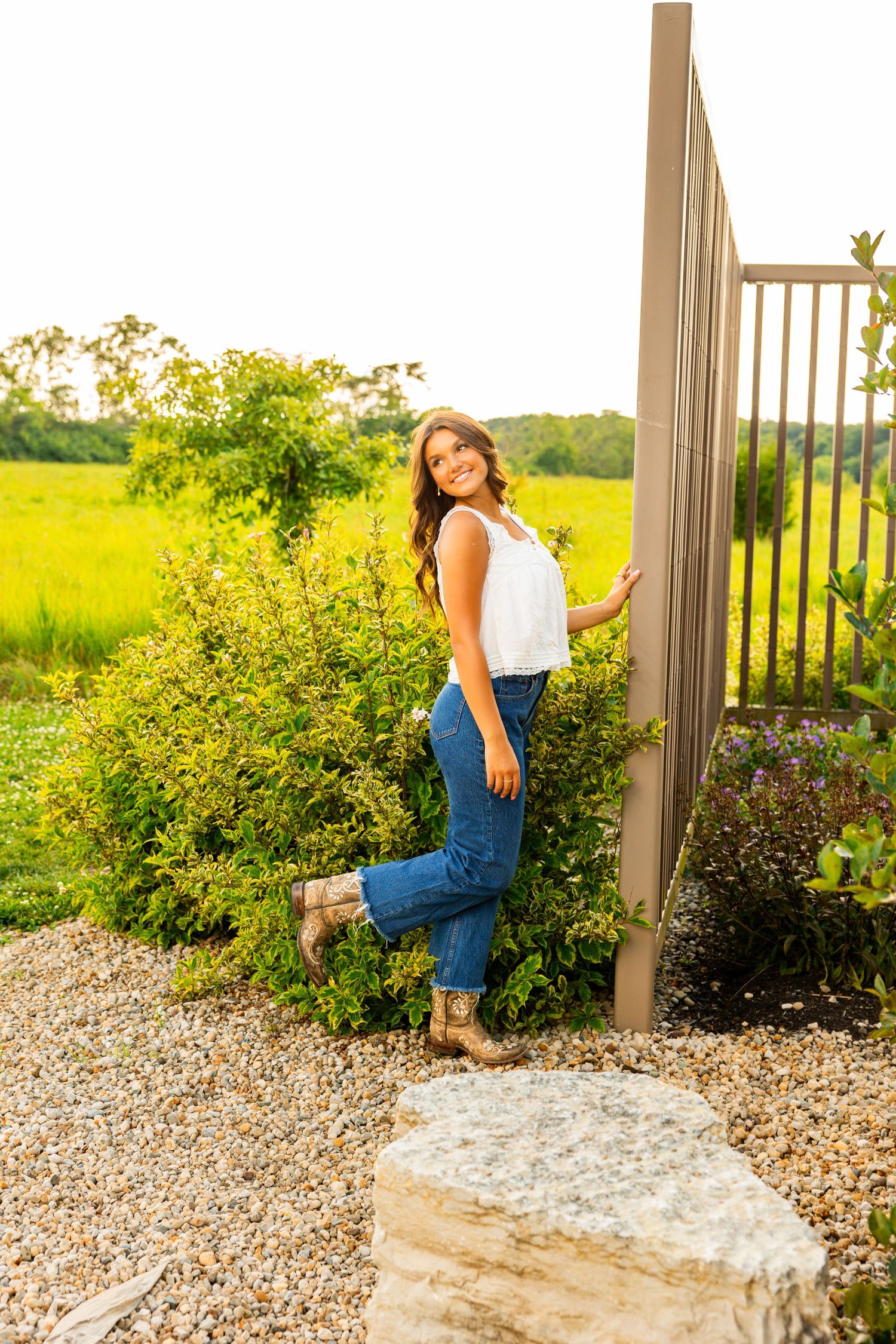 Young woman in white top and jeans poses by a garden fence with lush green fields in the background.