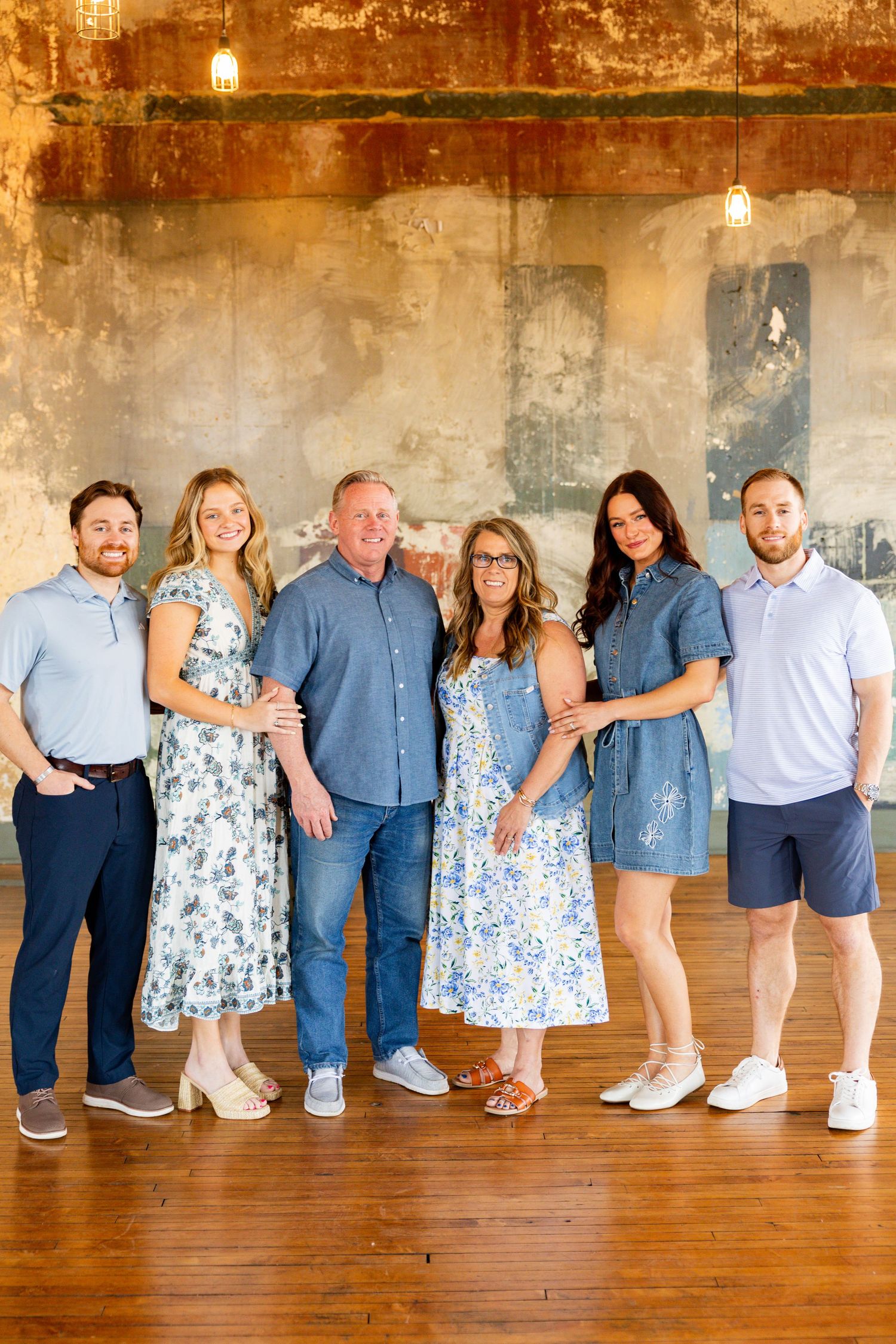 Six family members pose together smiling in a rustic indoor setting with warm wood floors and an aged wall backdrop.