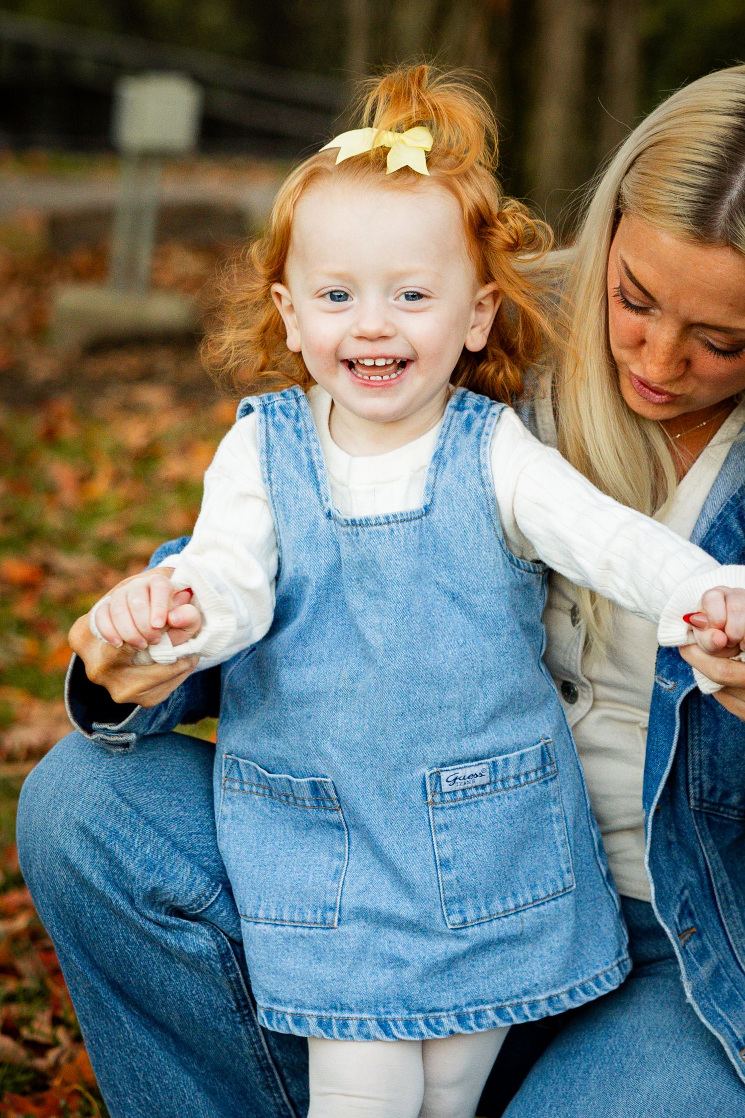 A small child wearing a denim jumper dress and white bow plays outdoors in autumn leaves.