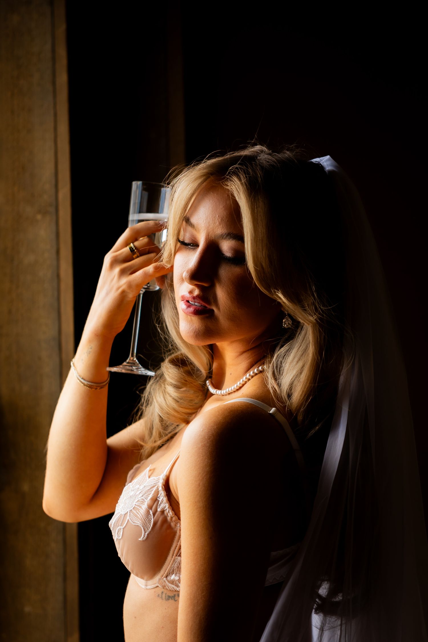 A woman in lingerie poses dramatically with a champagne glass near a window in moody lighting.