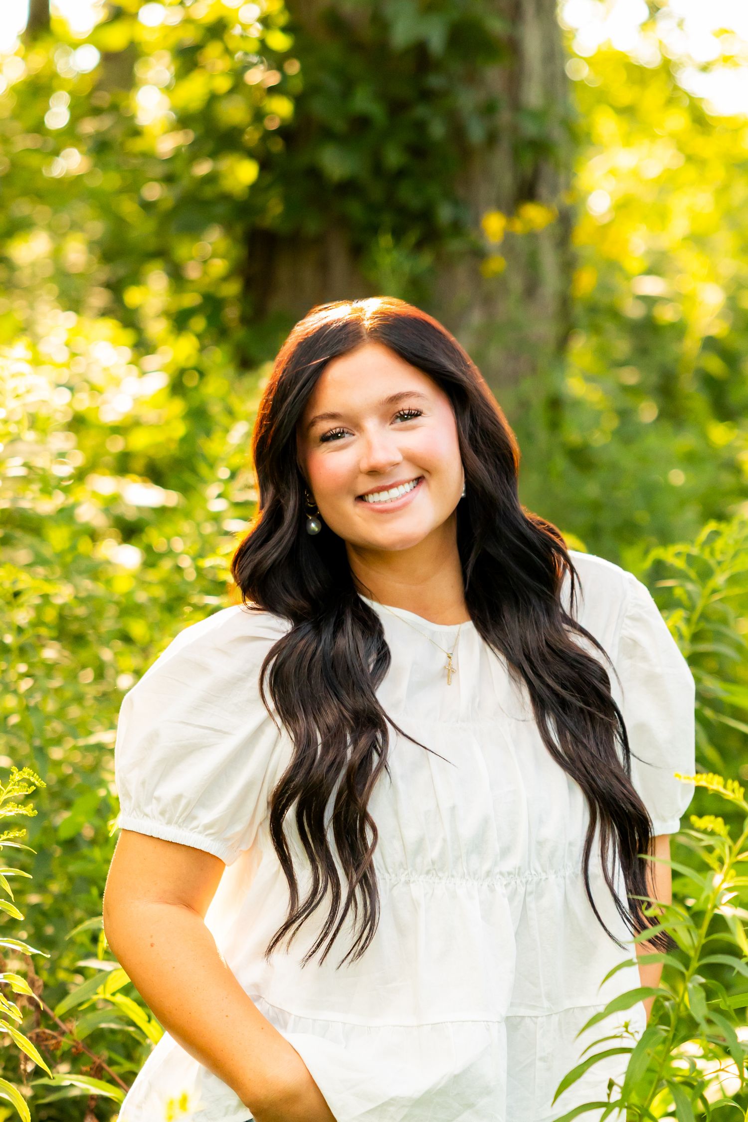 Young woman with long dark hair smiling in a white dress, posing outdoors among lush green foliage.