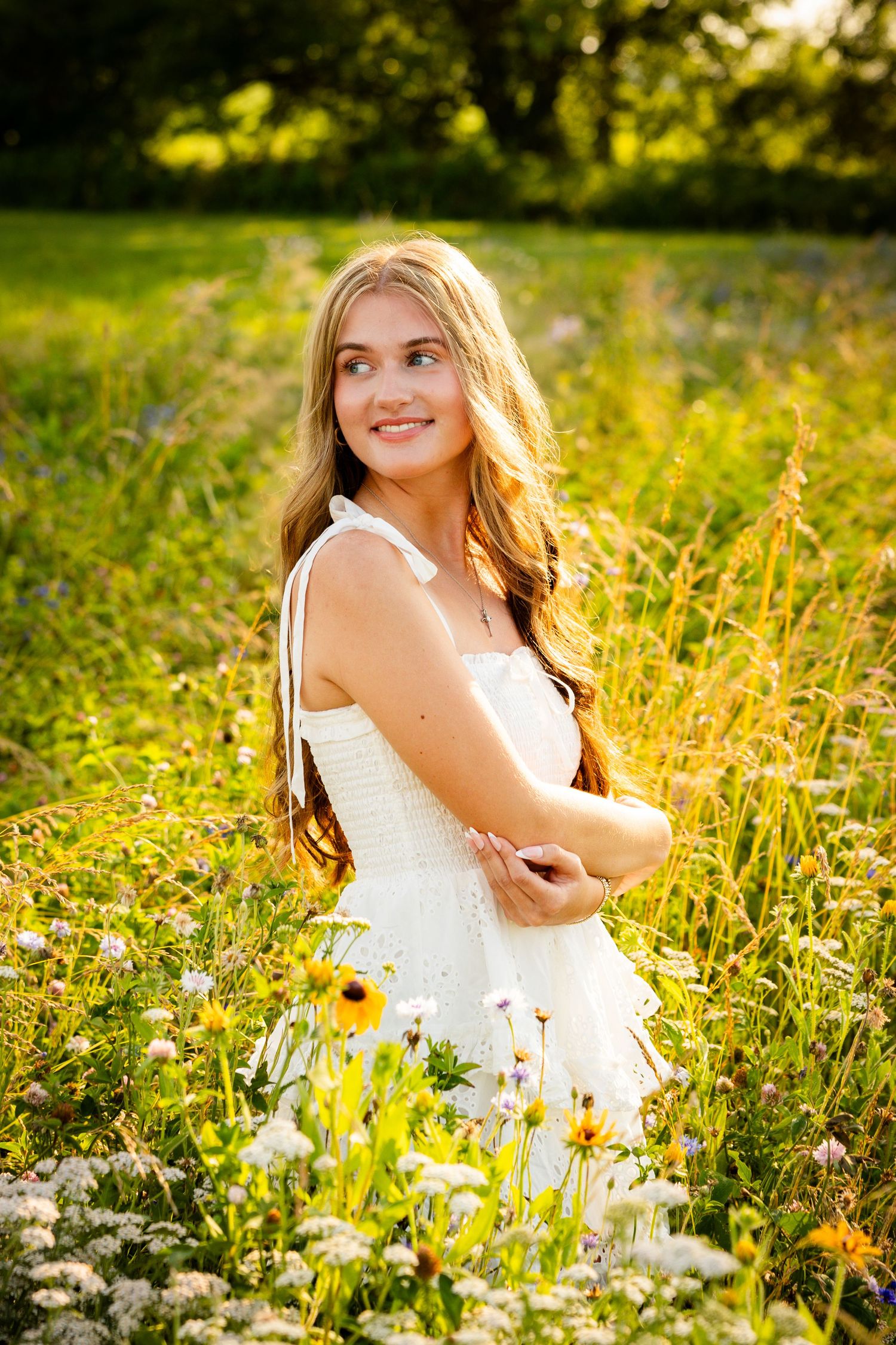 Young woman in white dress smiling in sunny wildflower meadow with golden light and green trees.