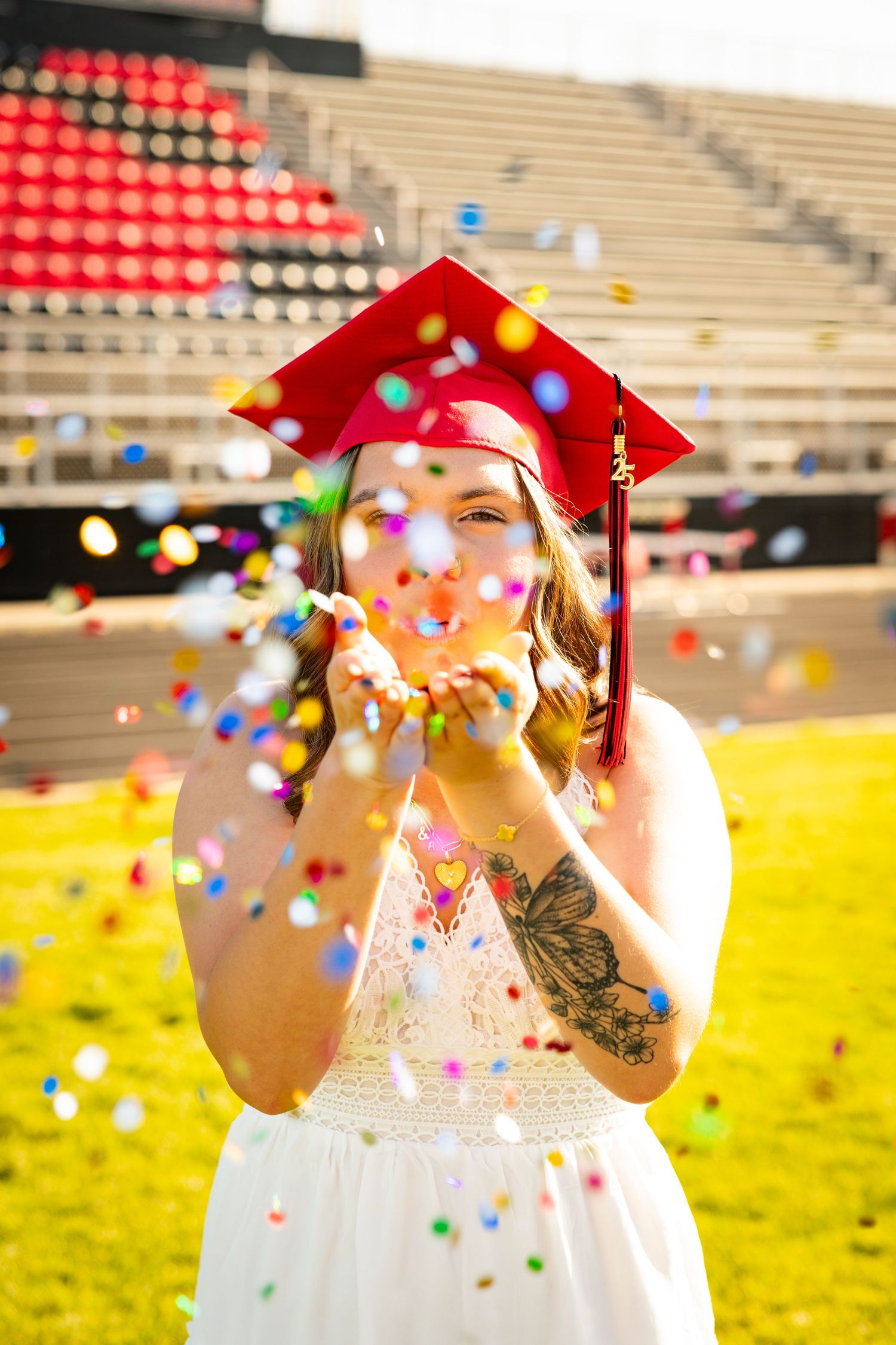 Graduate in white dress and red cap celebrates with colorful confetti falling on sunny grassy field near stadium stands.