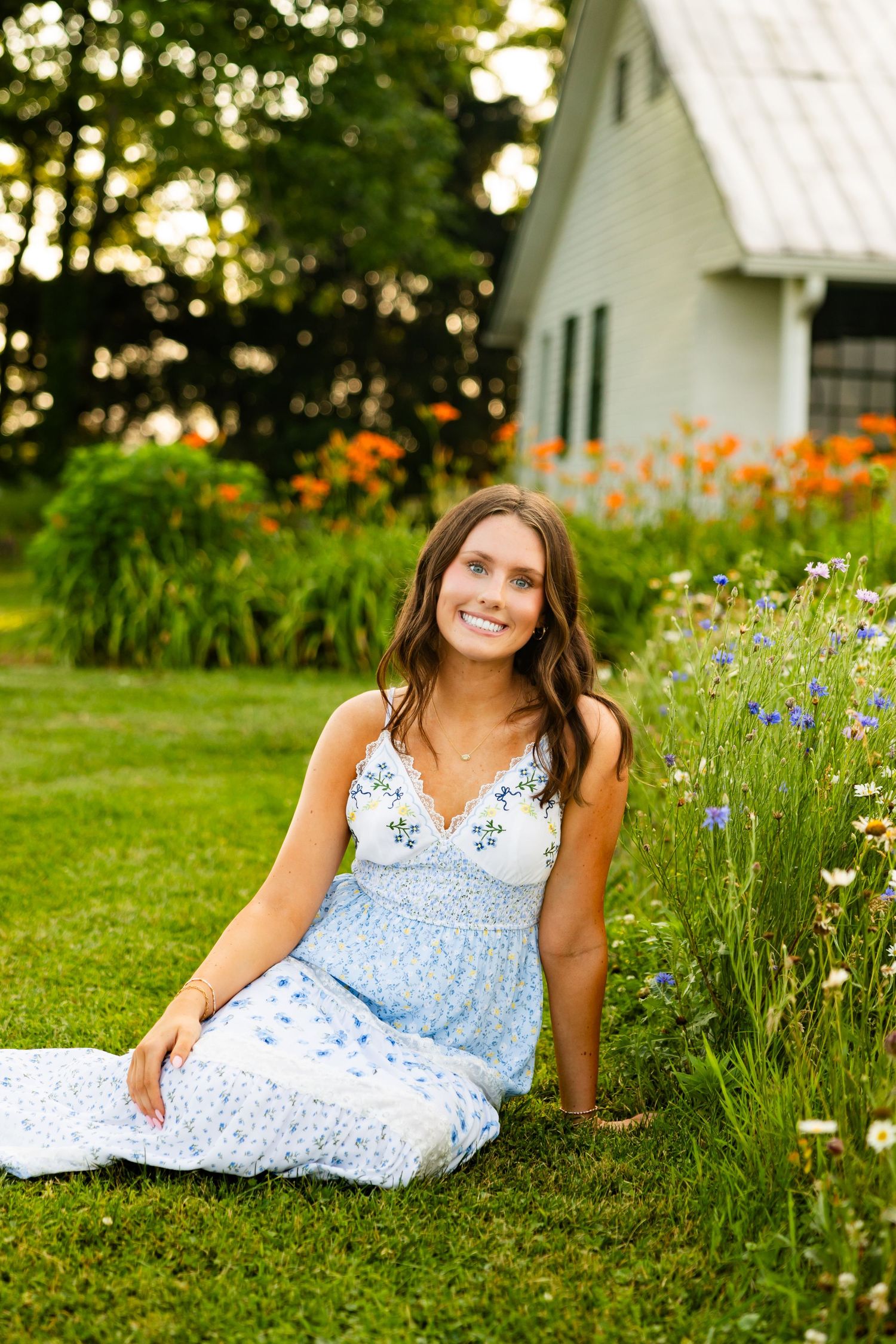 Young woman in blue floral dress sitting on grass beside wildflowers near a white cottage at sunset.