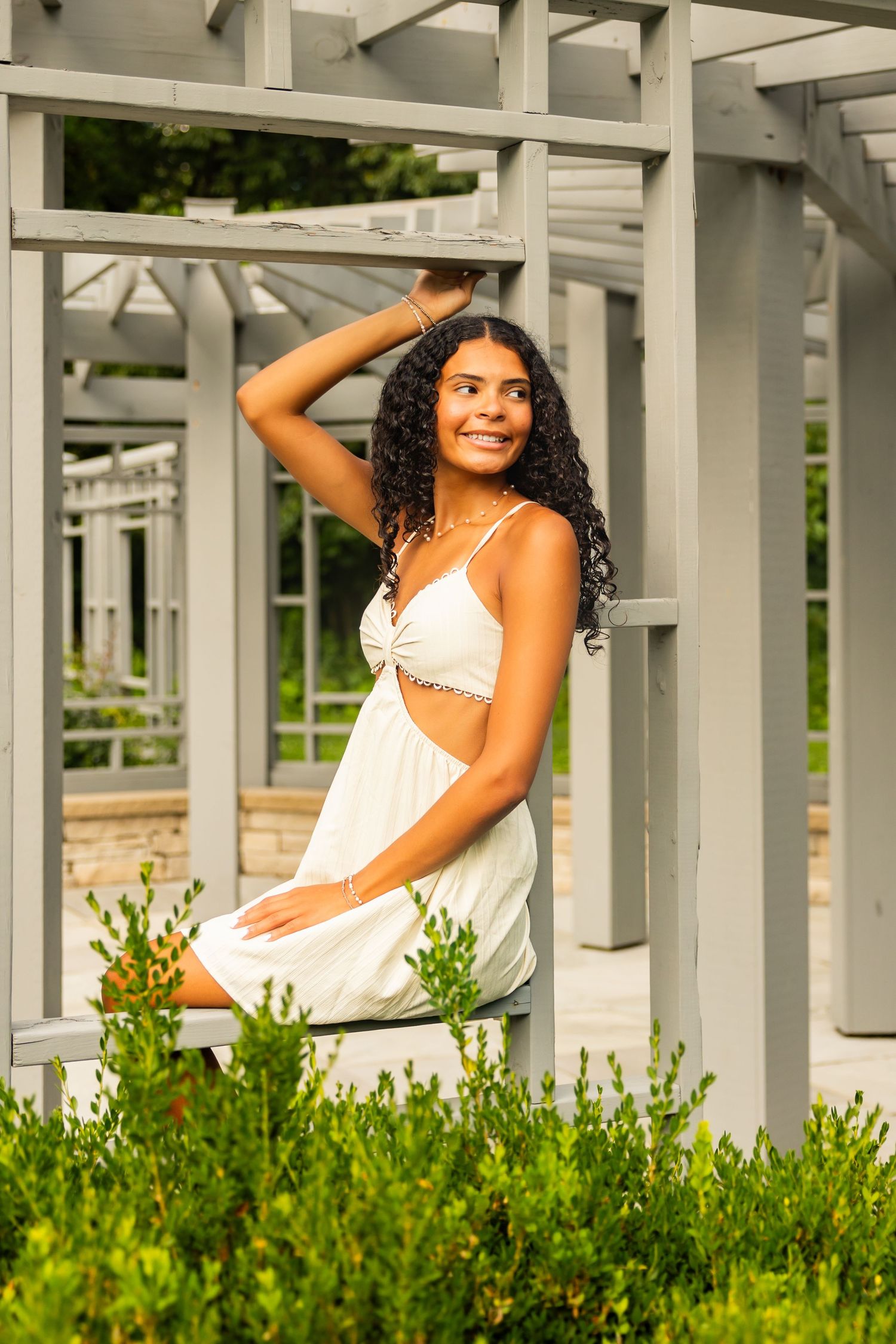 Young woman in white cutout dress poses gracefully under a pergola surrounded by lush green shrubs.