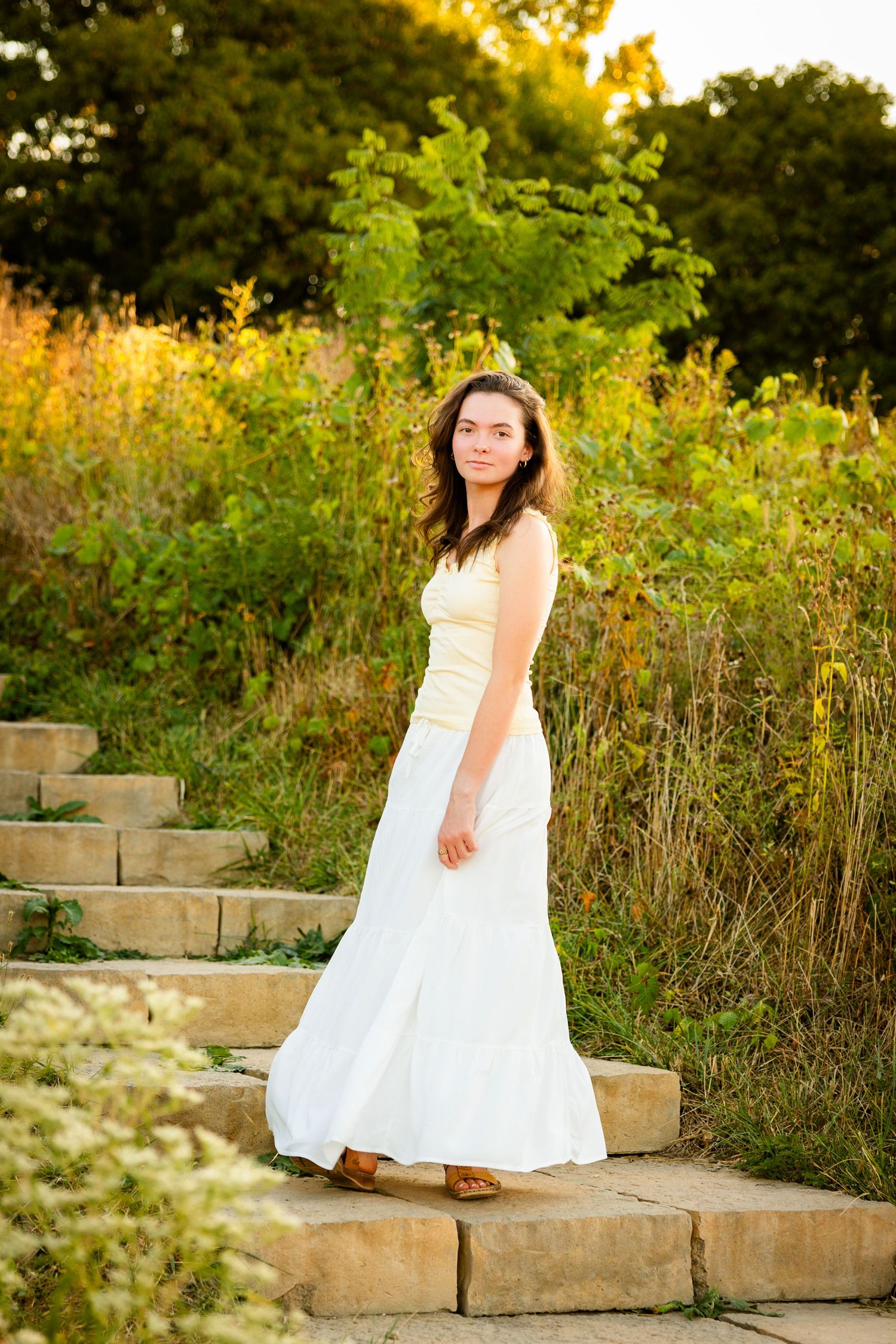 A person in a flowing white dress stands on stone steps at sunset surrounded by golden wildflowers and greenery.