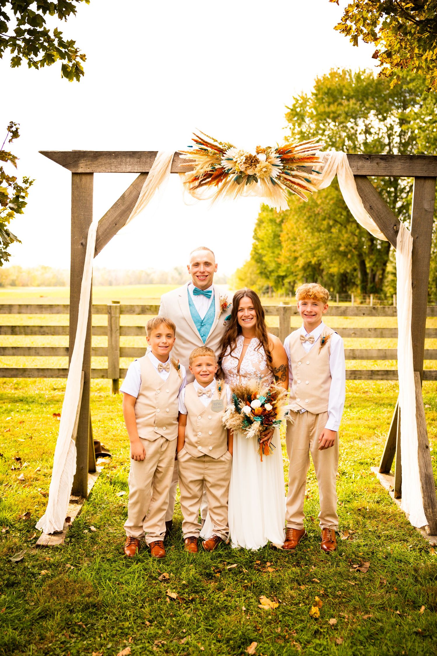 Wedding party stands in a row along rustic wooden fence at sunset in a grassy field with white fabric decor.