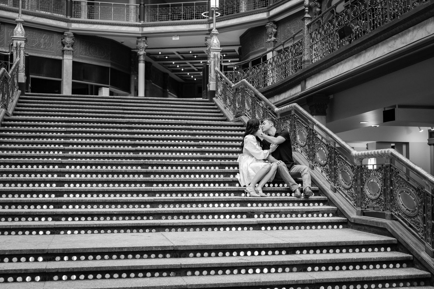 A couple shares an intimate moment on ornate grand staircase in black and white photography.