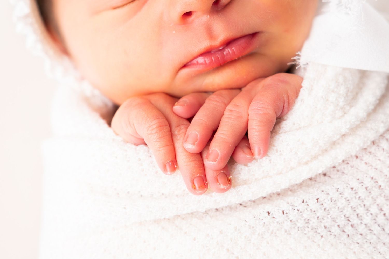 Close-up of a sleeping newborn baby swaddled in white, with tiny delicate fingers resting near their chin.