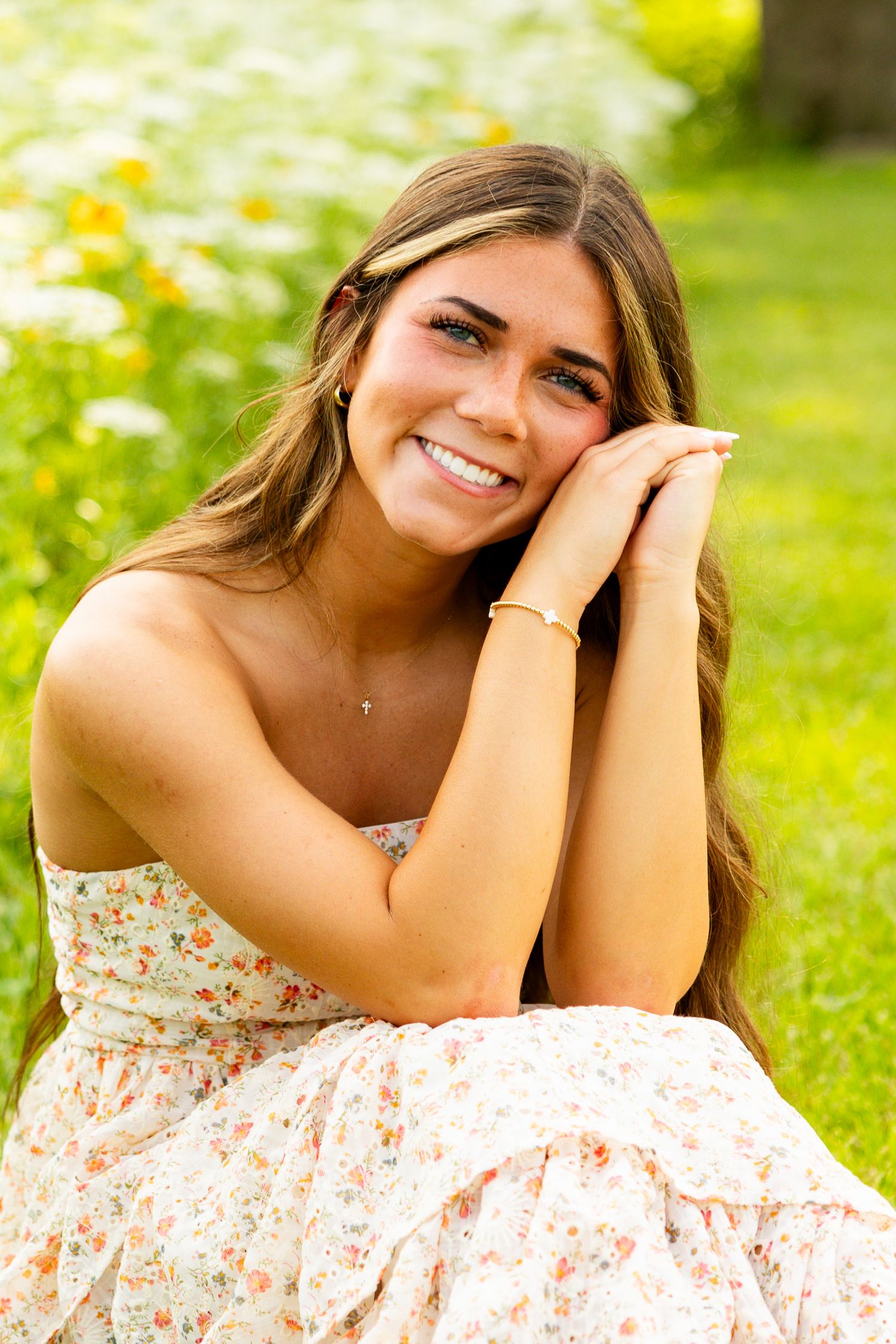 Smiling young woman in floral sundress sitting outdoors in a garden with wildflowers, resting her chin on her hands.