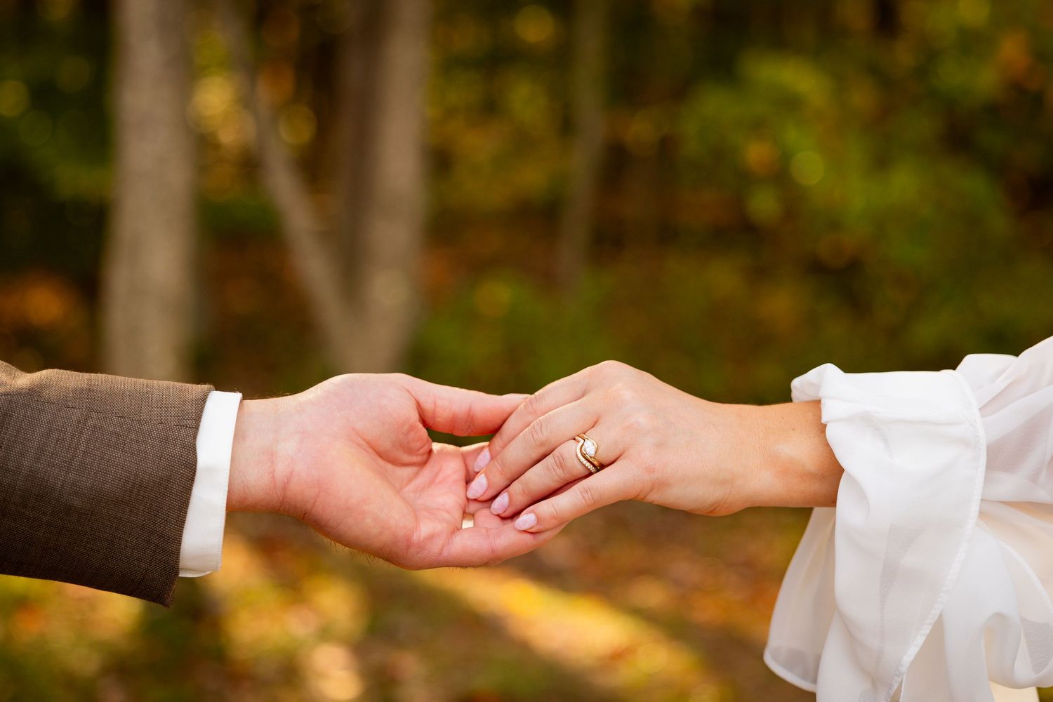 Two hands tenderly touch during a romantic autumn wedding ceremony with blurred fall foliage in background.