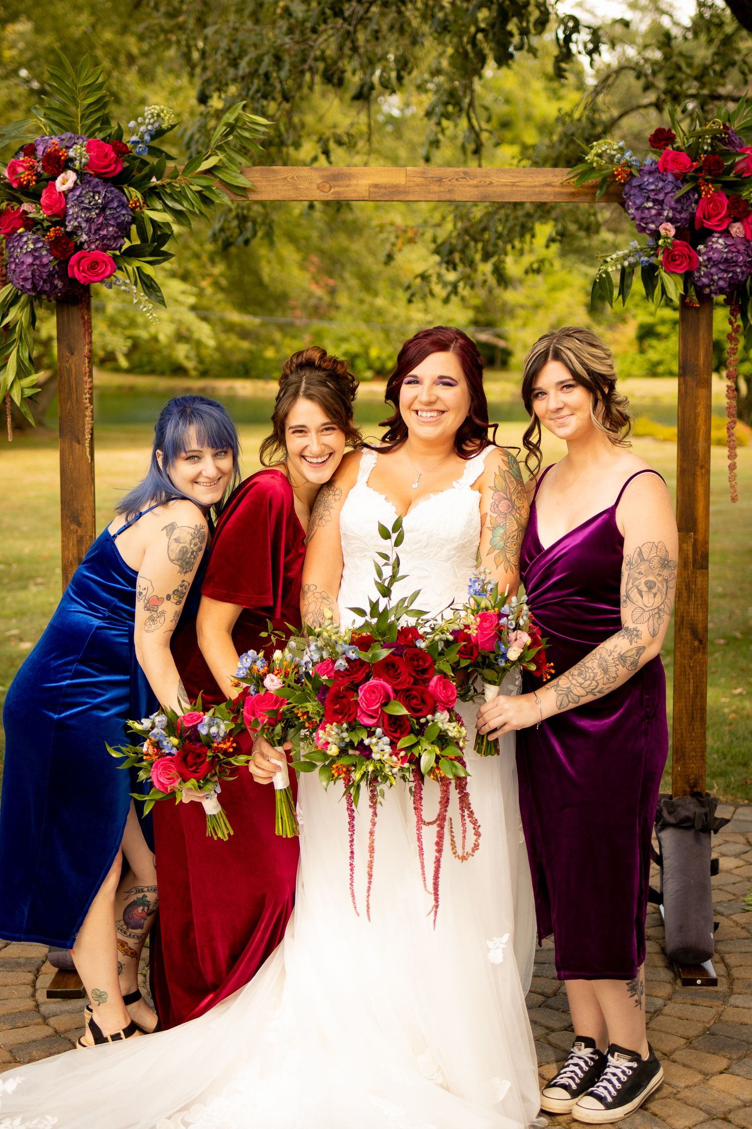 A wedding party poses outdoors wearing burgundy and royal blue dresses with vibrant red floral bouquets.