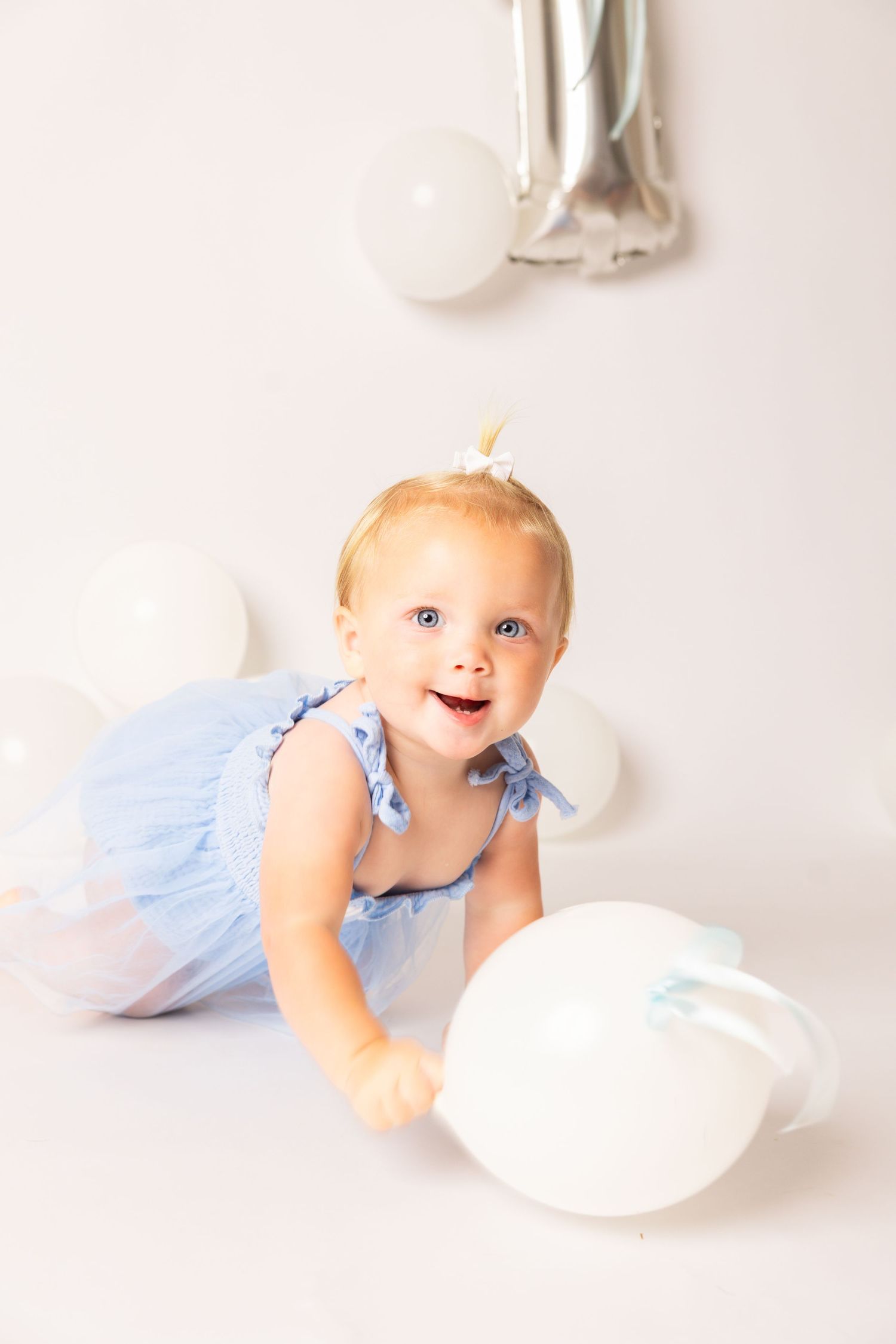 Smiling baby girl in a blue dress crawls toward a white balloon during a first birthday photo shoot with a silver number one balloon.
