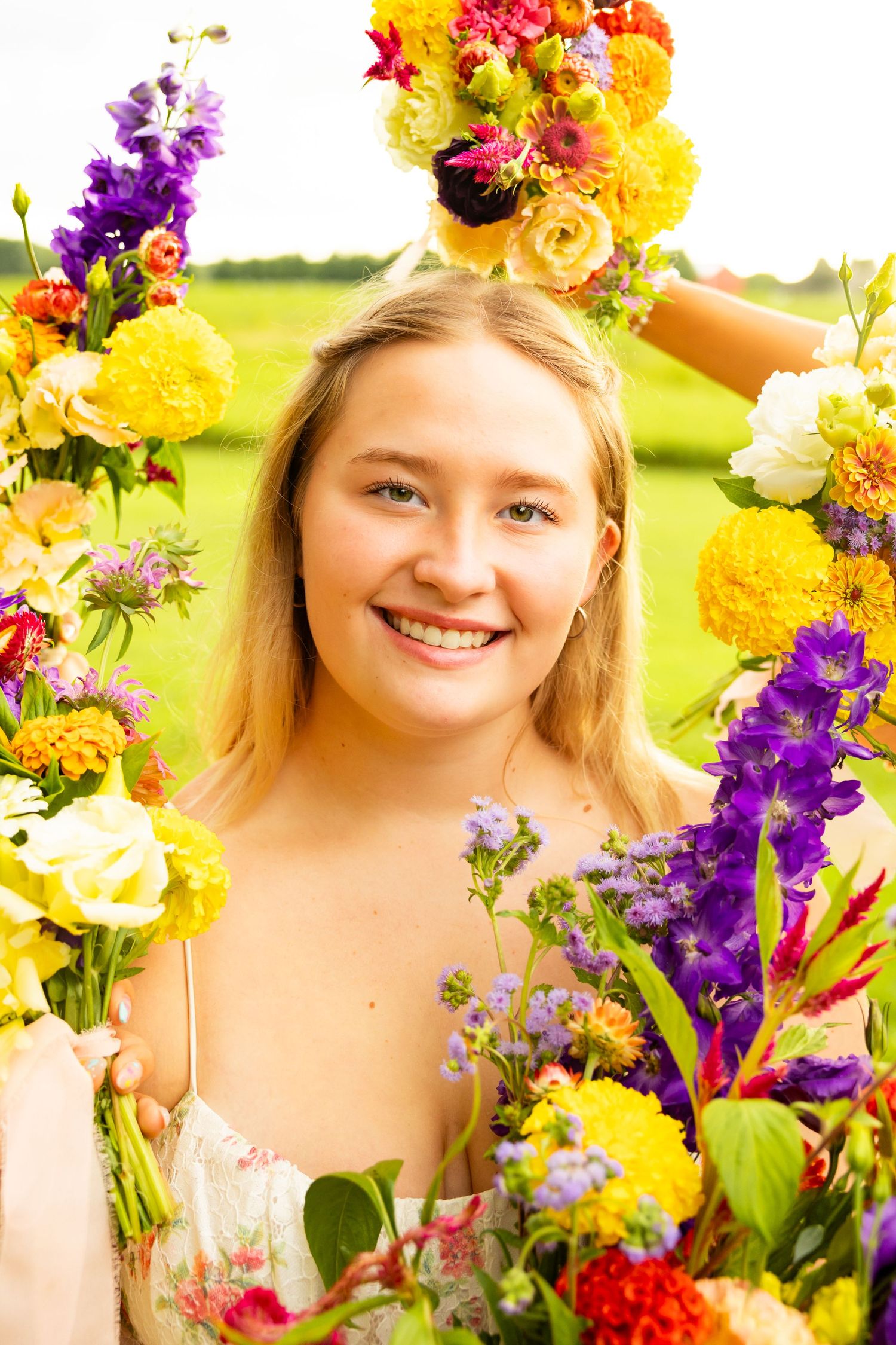 Smiling young blonde woman surrounded by colorful yellow, purple, and pink flowers outdoors.