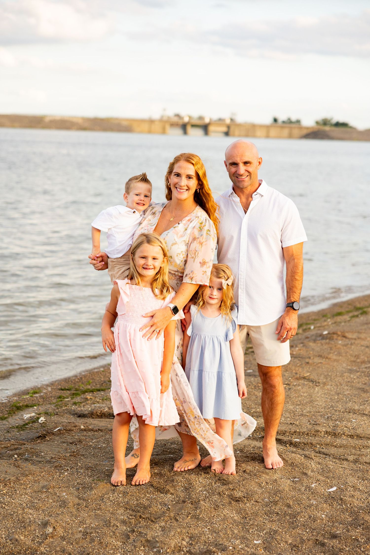 Smiling family of five in light summer outfits posing barefoot on a sandy lakeshore at golden hour.