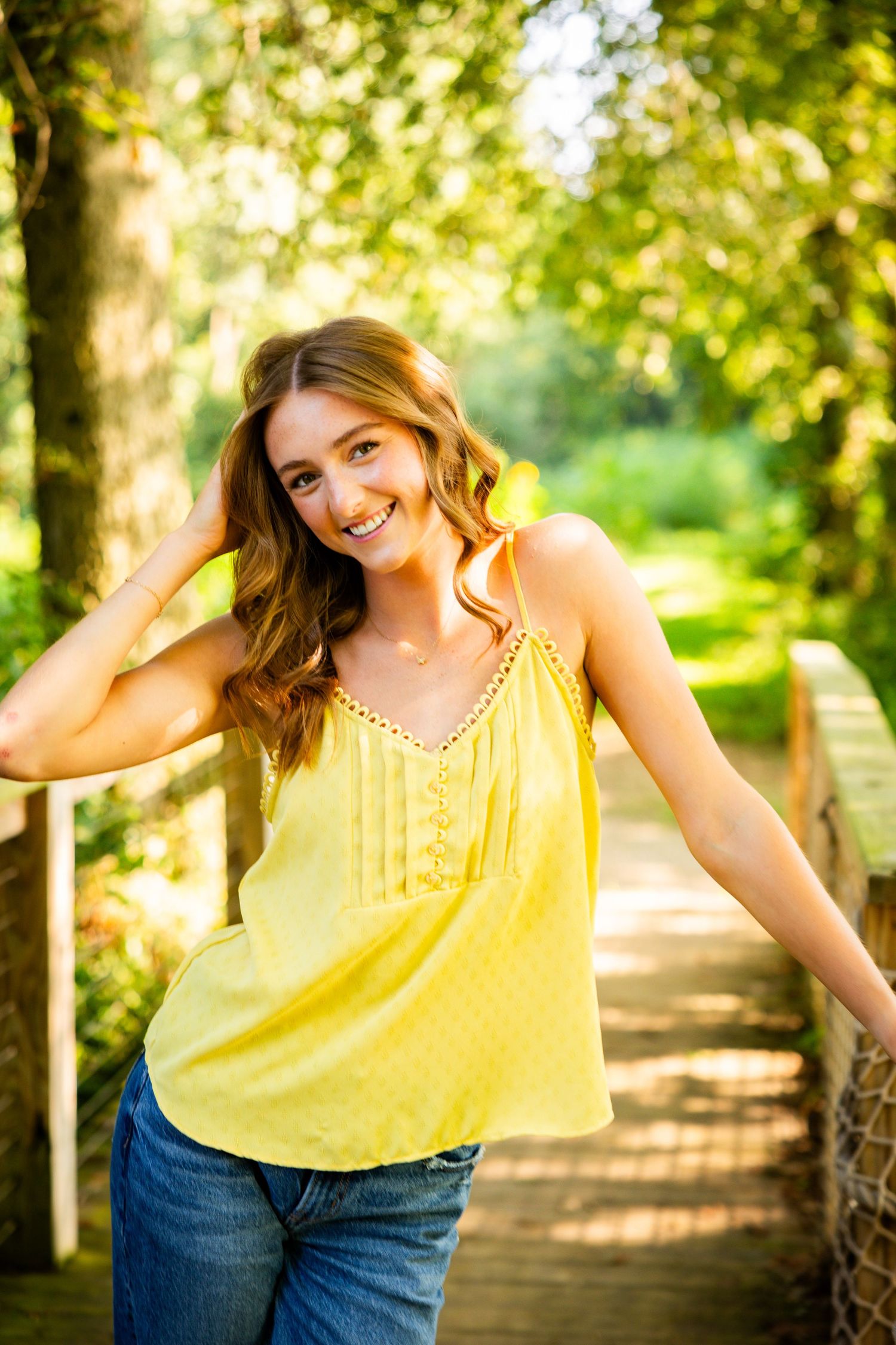 A person in a yellow sleeveless top and jeans poses playfully on a sunlit wooden path through trees.