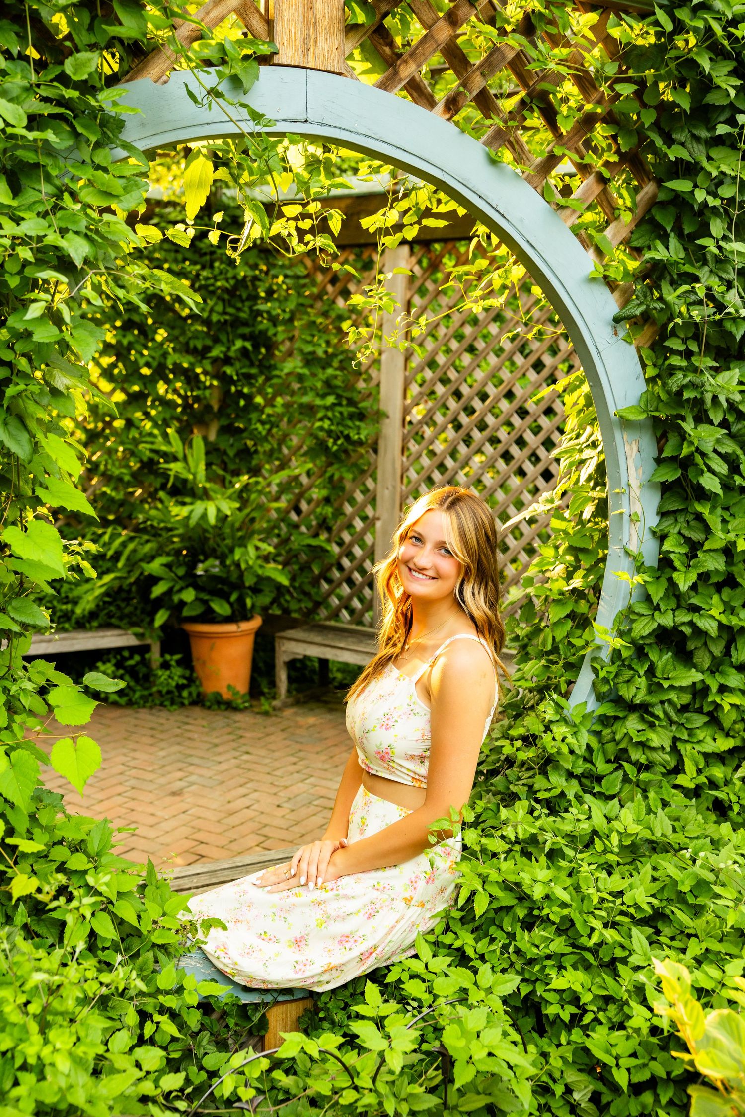 Young woman in floral dress smiling inside a circular garden arch surrounded by lush green ivy and wooden lattice.