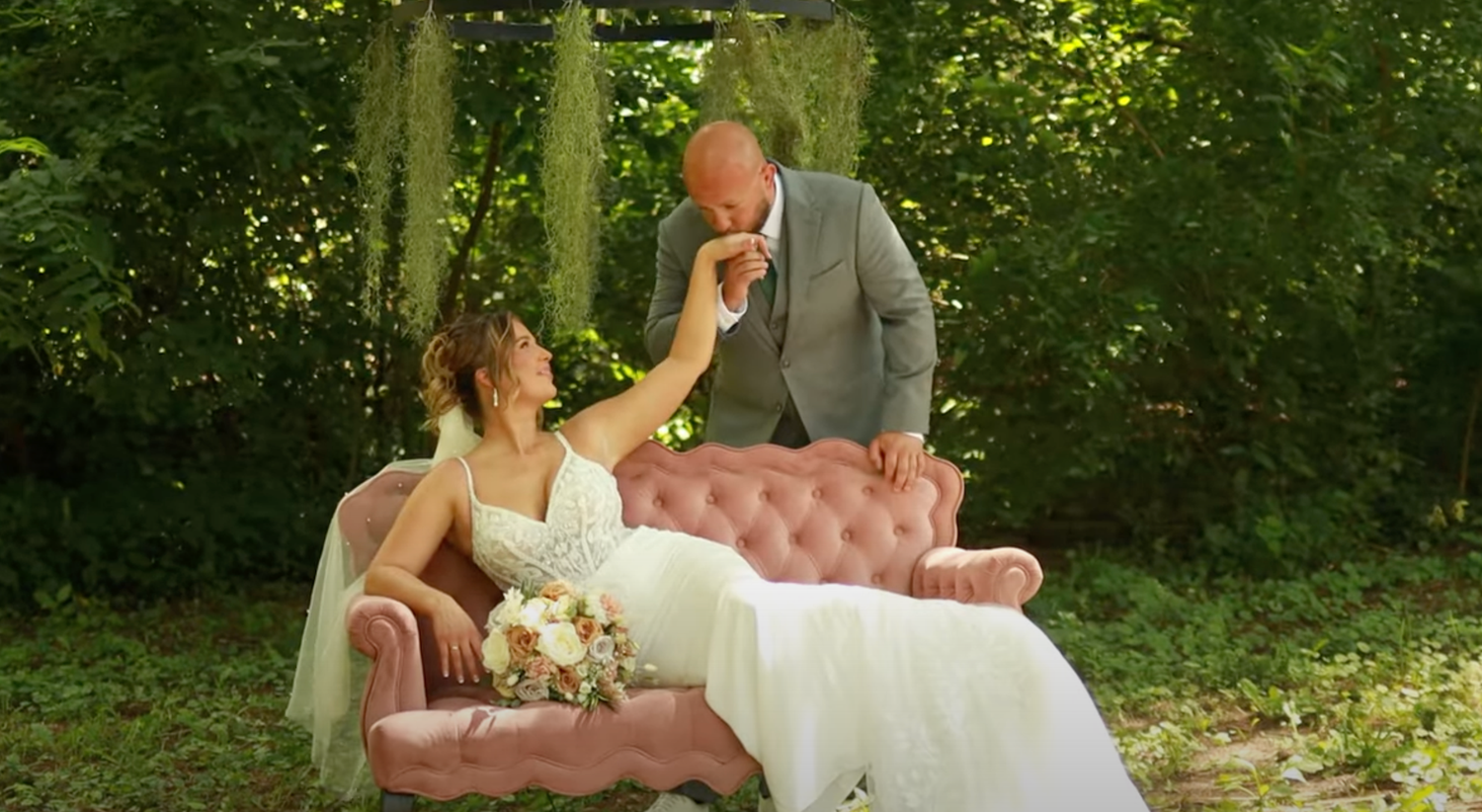 A couple shares a romantic moment on a pink vintage couch outdoors in a lush garden setting with a white floral bouquet.