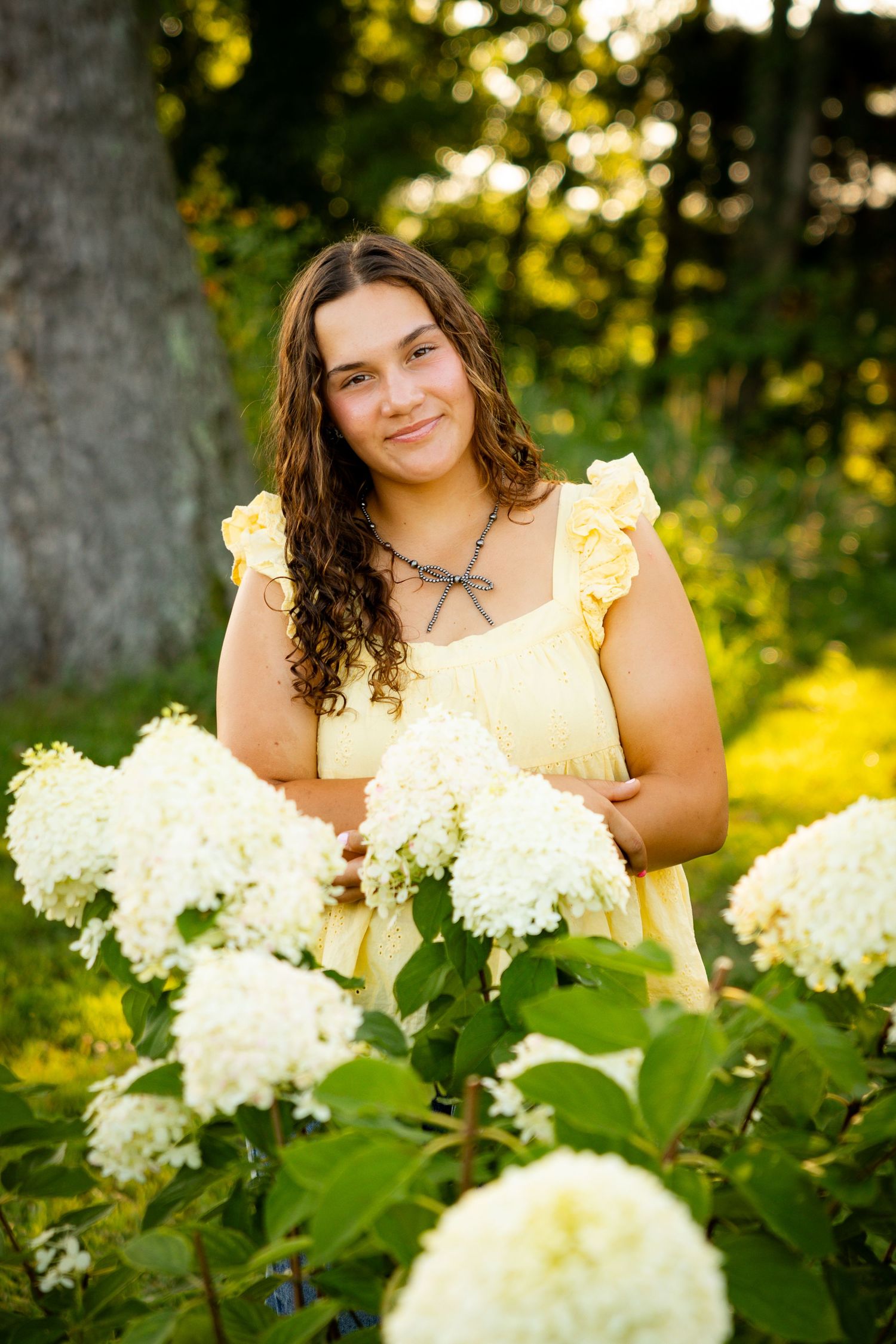 A woman in a yellow dress poses near blooming white hydrangea flowers in a garden during golden hour.