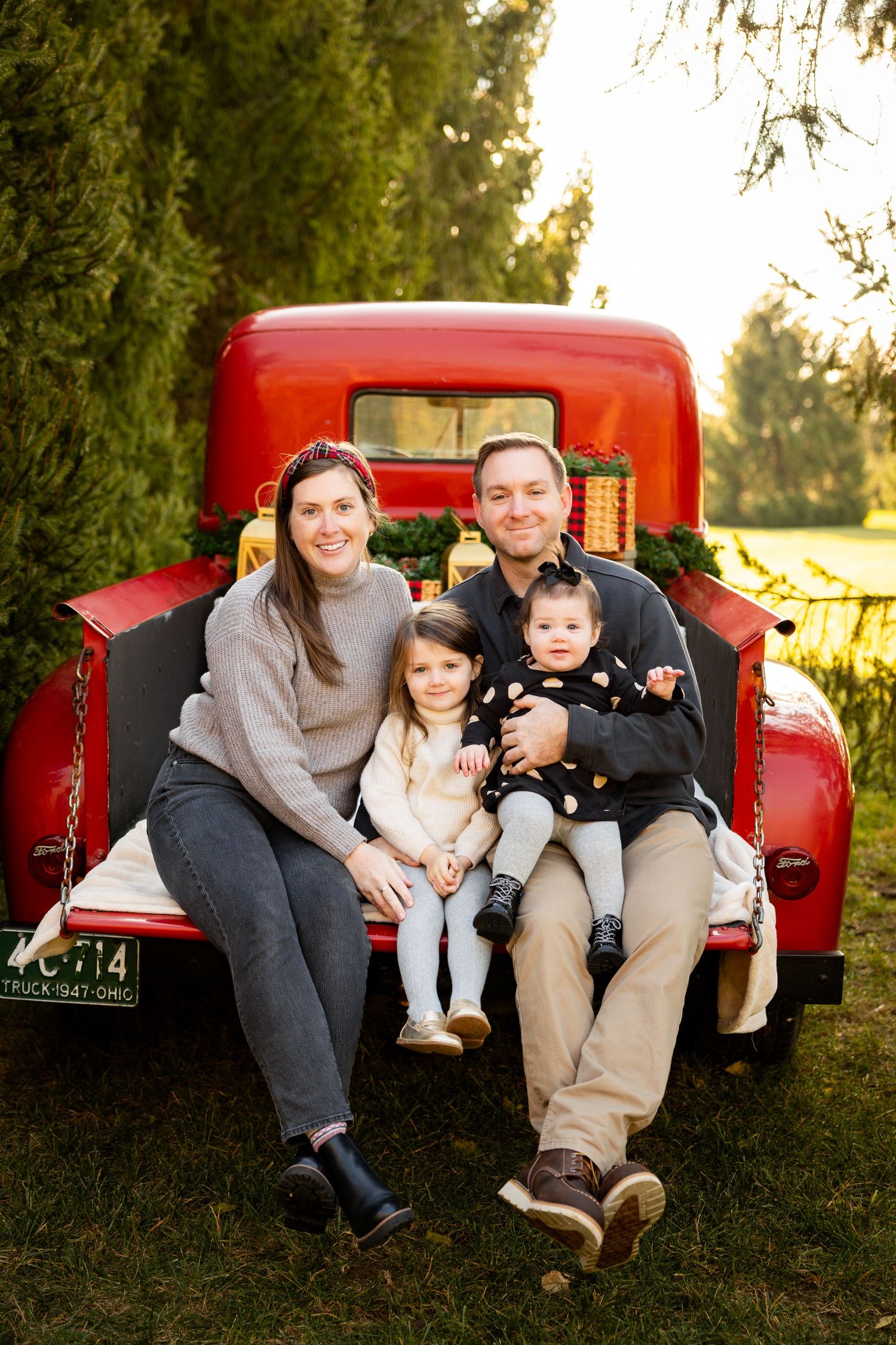 A family sits together on the tailgate of a vintage red pickup truck during a sunny outdoor photo session.