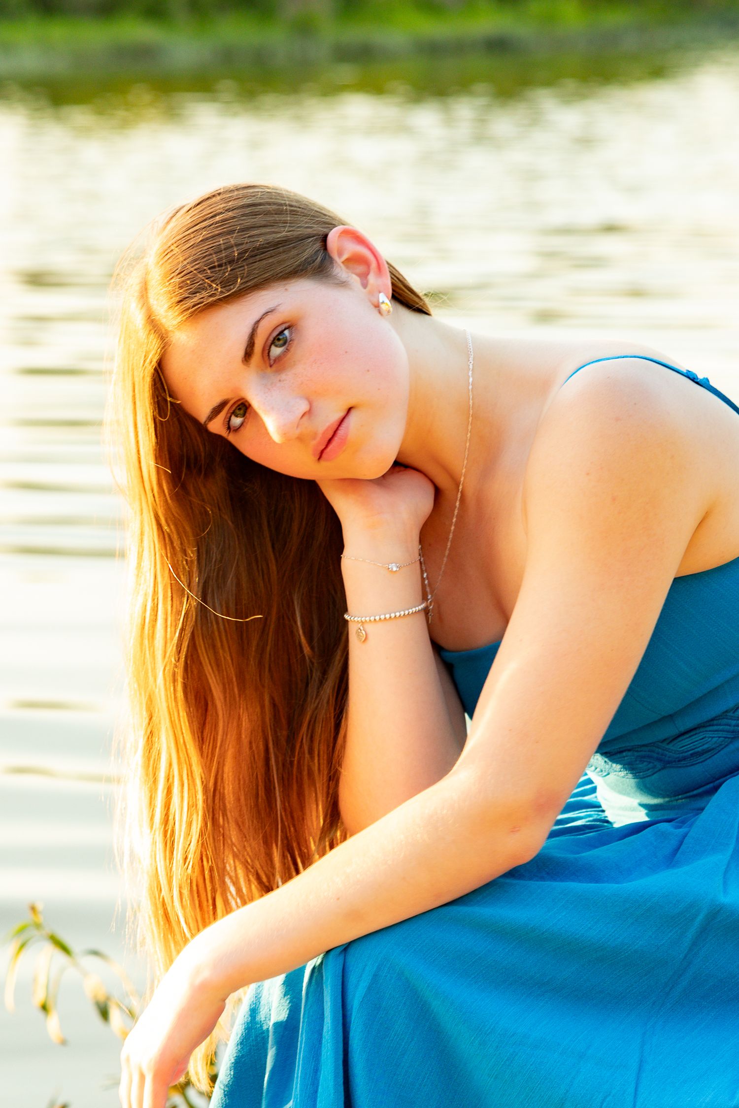 Young woman with long auburn hair in a teal dress sits by a reflective lake in golden hour light.