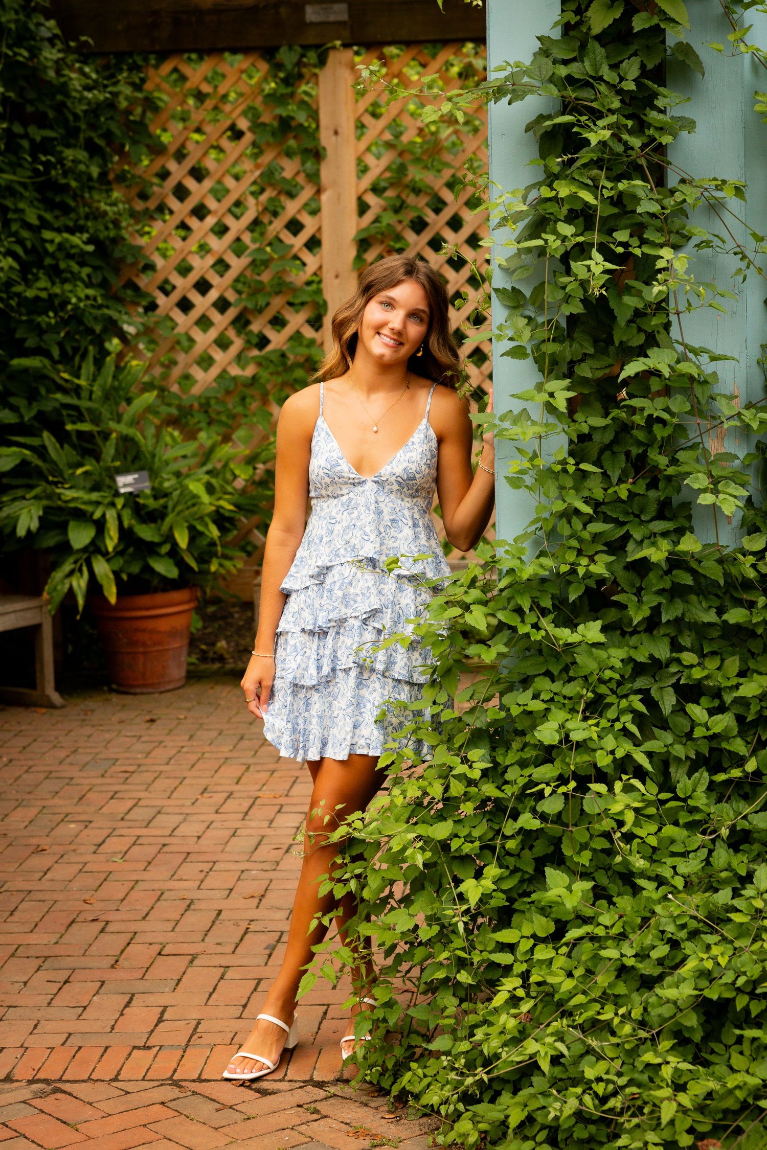 Woman in a floral summer dress poses in front of a garden trellis with climbing vines on a brick patio.