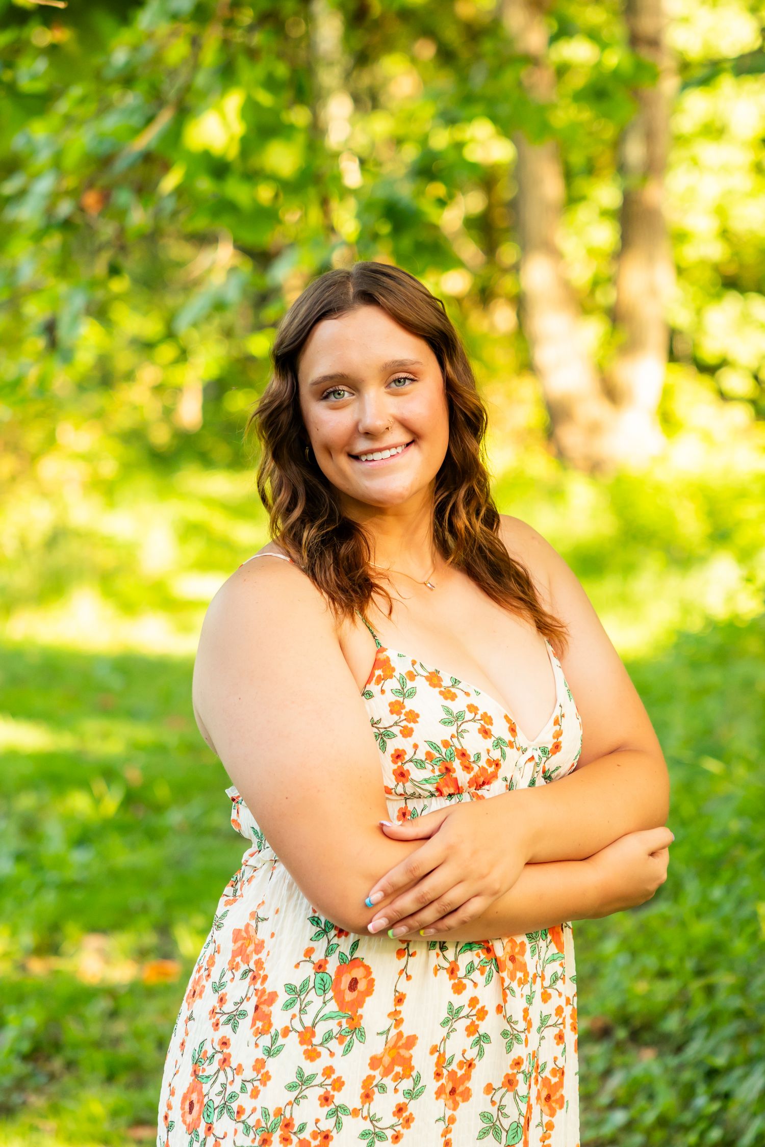 Smiling young woman in floral sundress with arms crossed, posing outdoors in a lush green park setting.