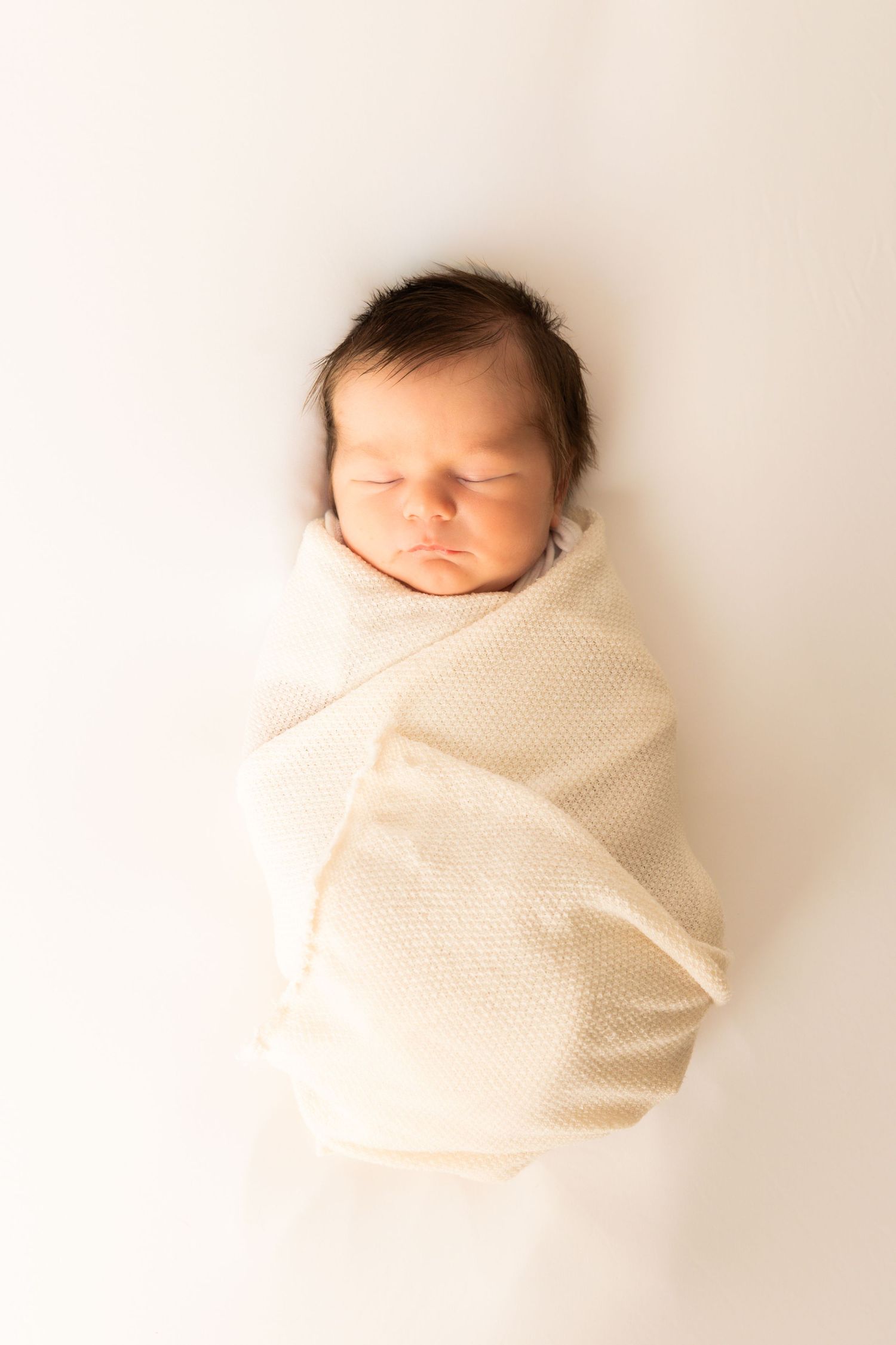 Sleeping newborn baby with dark hair swaddled in a cream knit blanket against a soft white background in a peaceful newborn portrait.