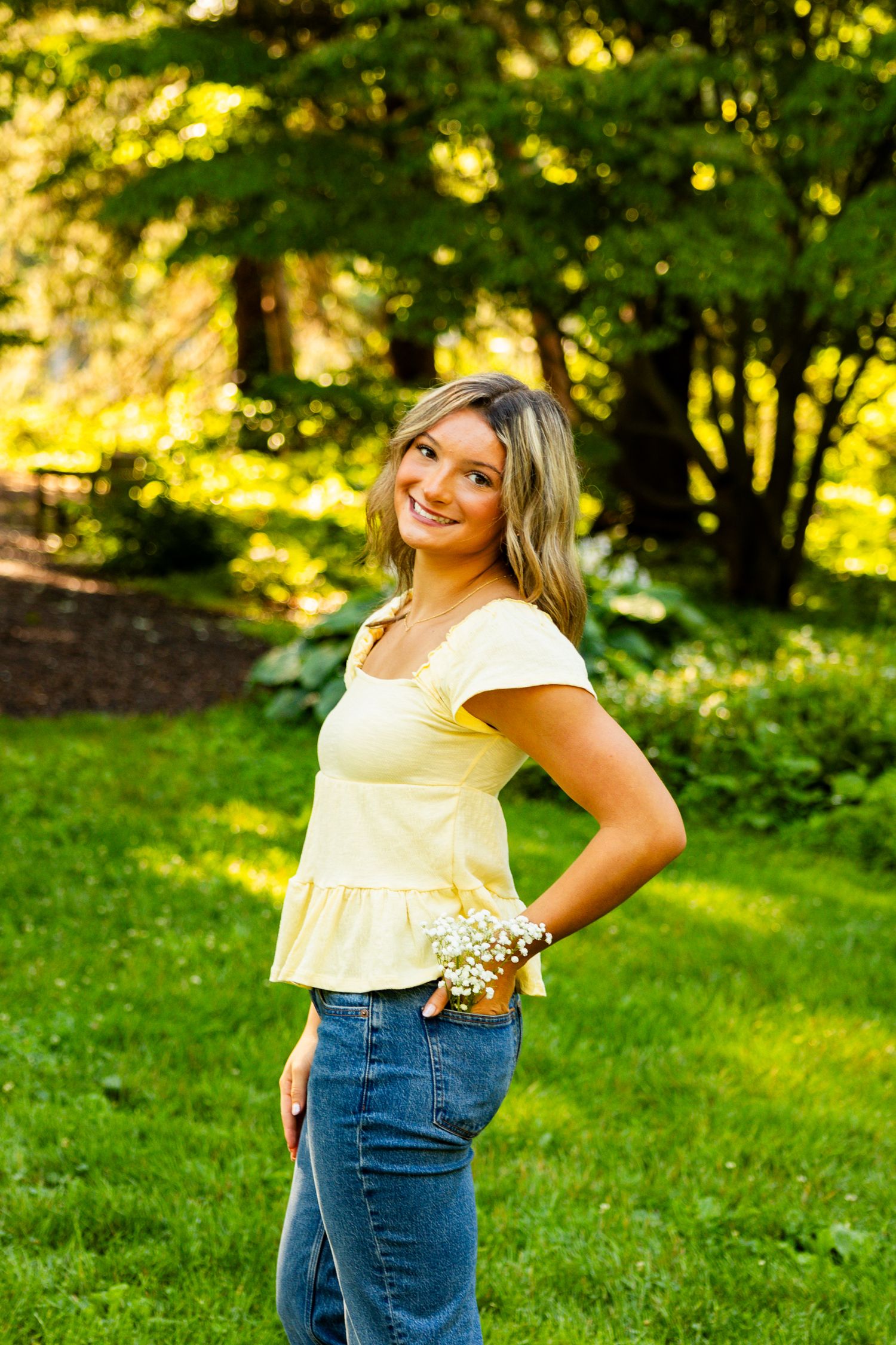 Young woman in yellow top and jeans smiling outdoors in a lush green garden setting.