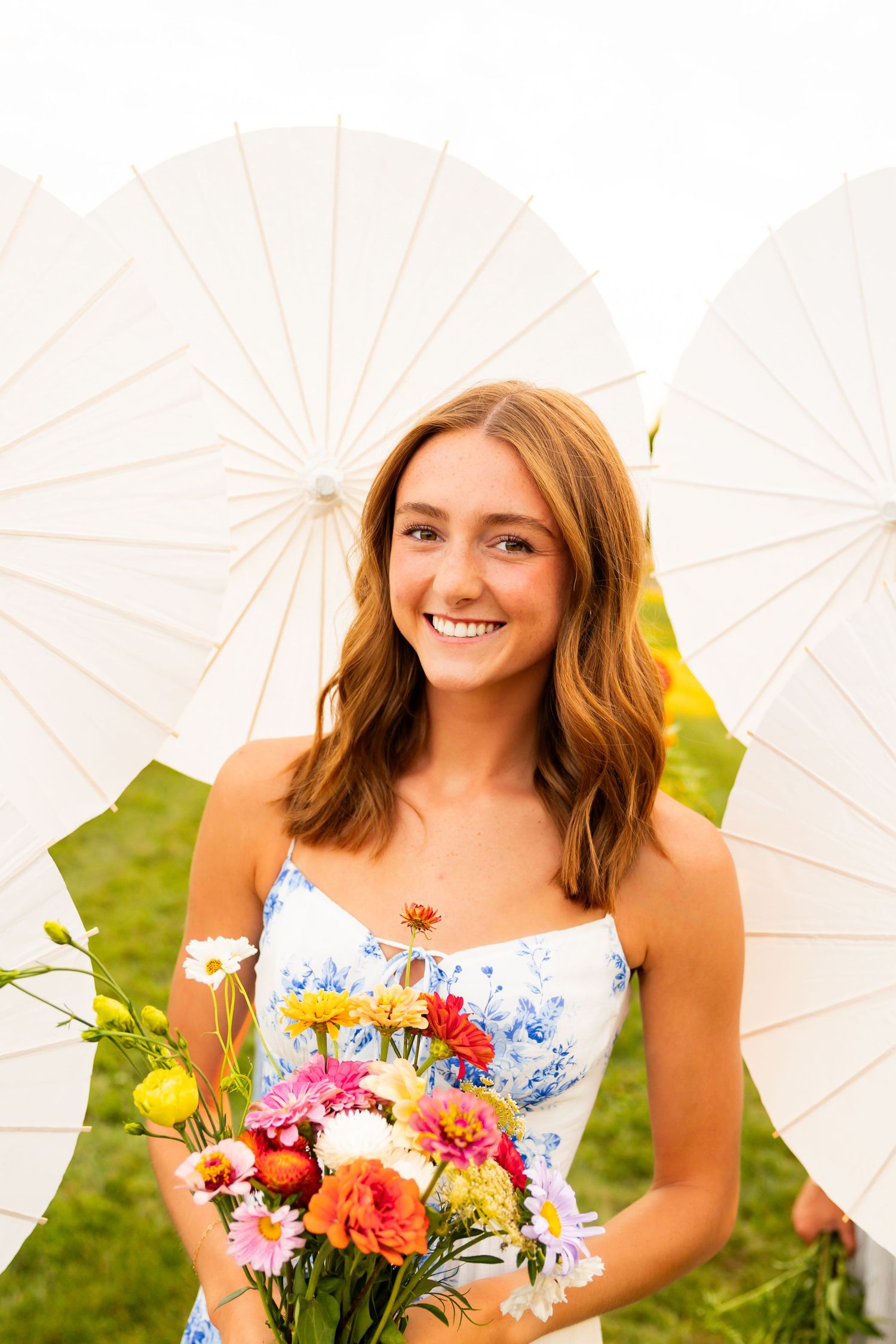 Person in white floral dress holds colorful bouquet of flowers while posing with white parasol on sunny lawn.