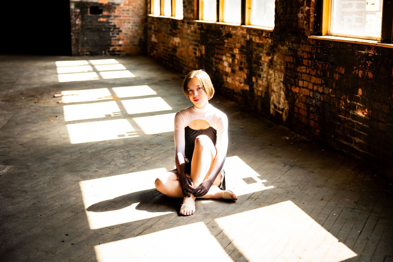 A dancer in black leotard strikes graceful poses in a sunlit industrial loft space with brick walls and wooden floors.