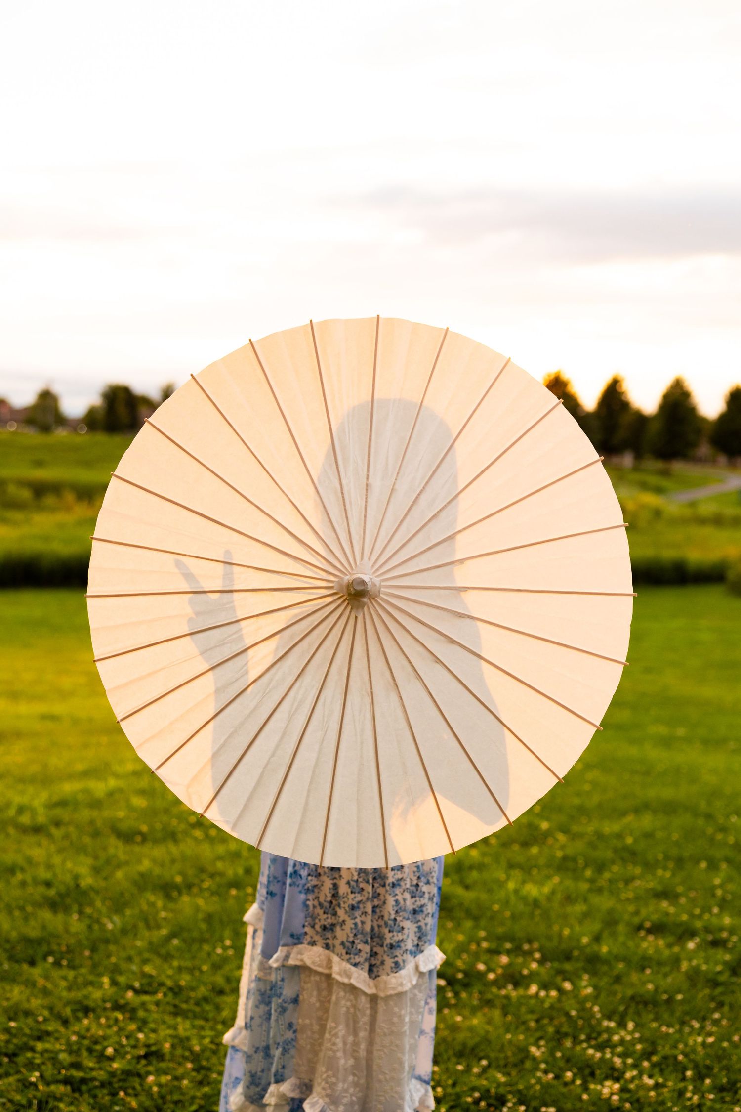 Person holding a white paper parasol umbrella in a green field at sunset, wearing a floral dress.