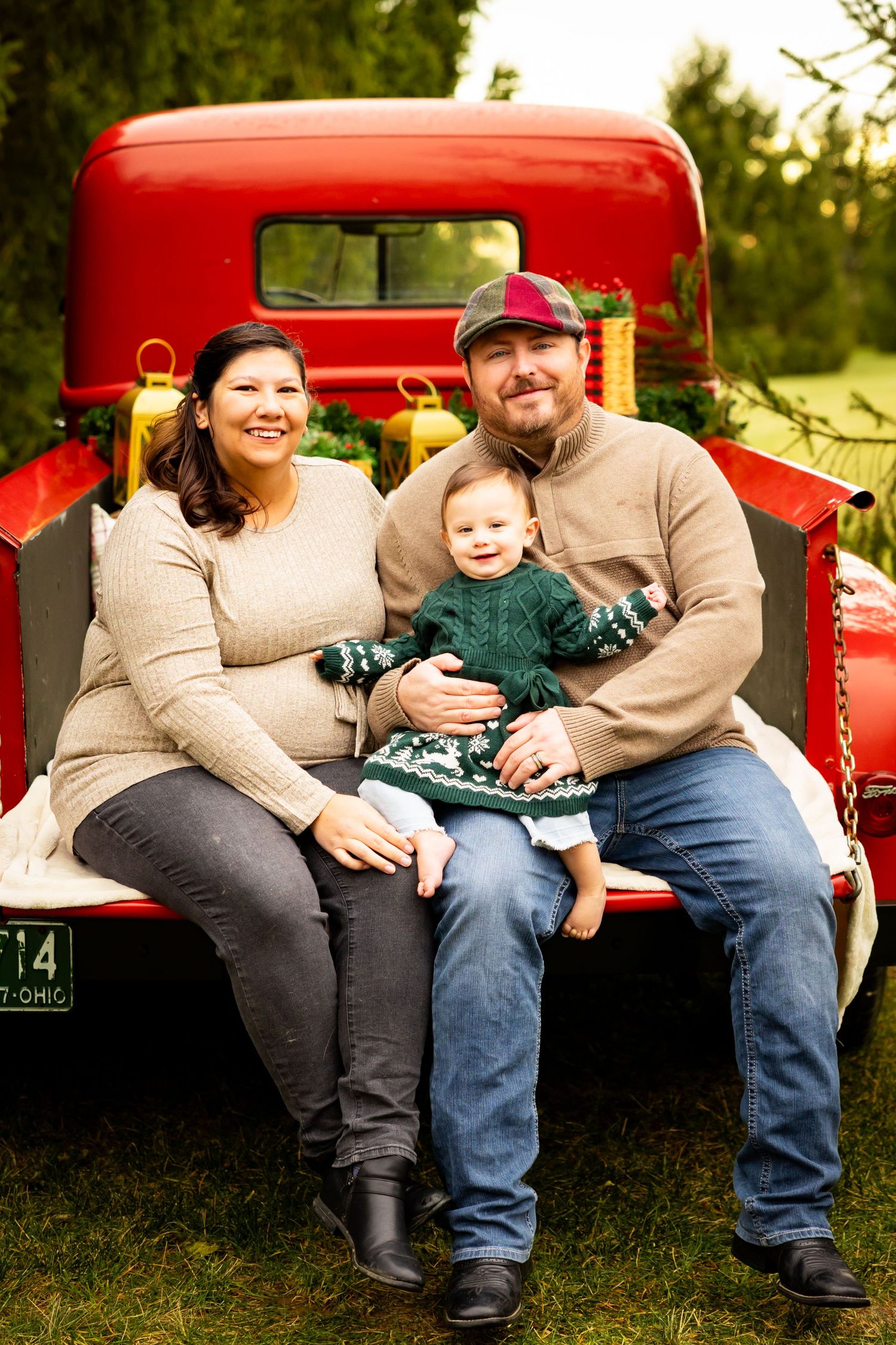 A family sits together on the tailgate of a vintage red truck during a sunset photo session in a grassy field.